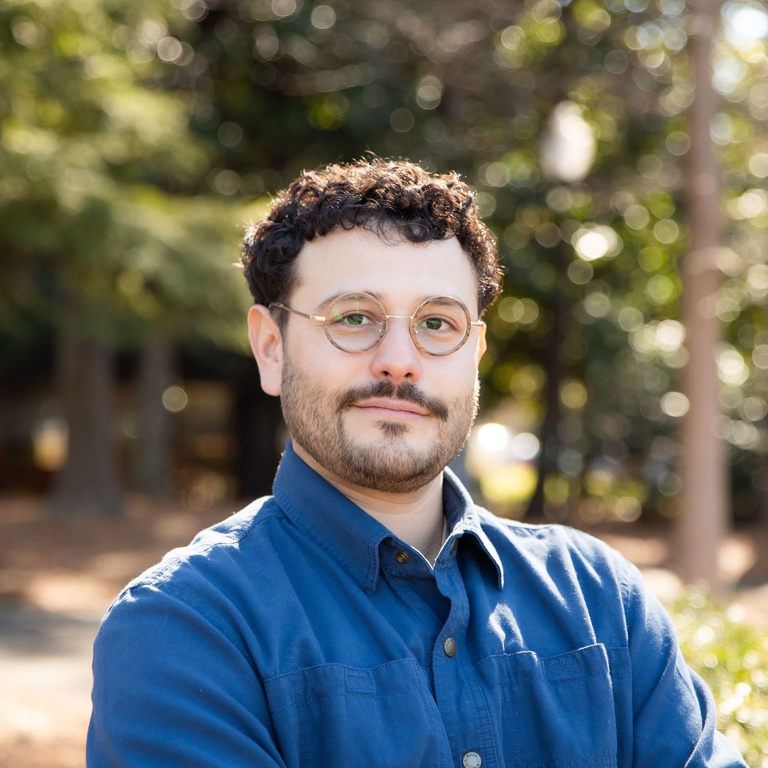 Professional headshot of a person in a blue shirt against a blurred outdoor background with trees.