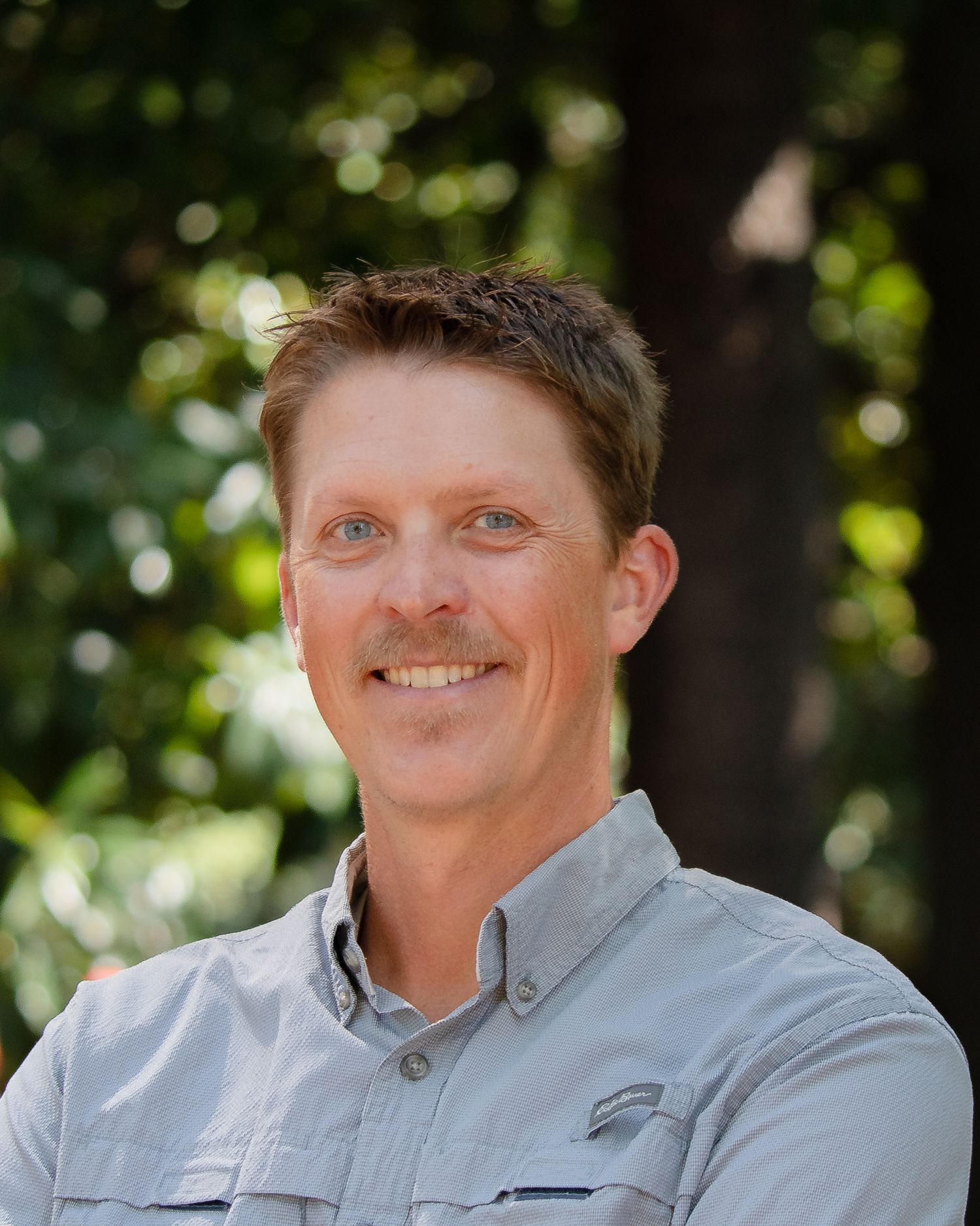Professional headshot of a person in a light gray button-down shirt smiling outdoors with blurred foliage in background.