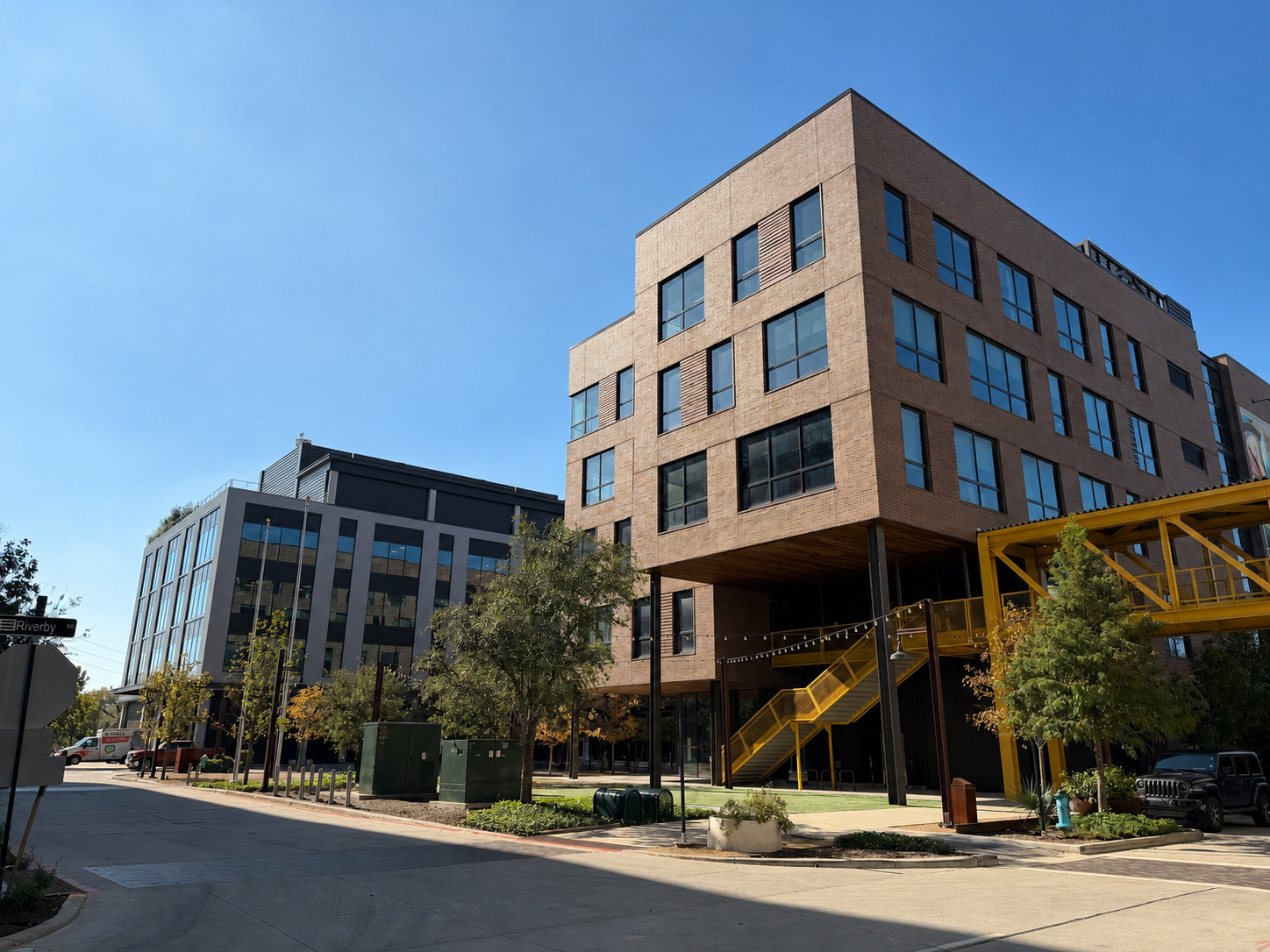 Modern brick office buildings with yellow staircases along a sunny urban street lined with trees.