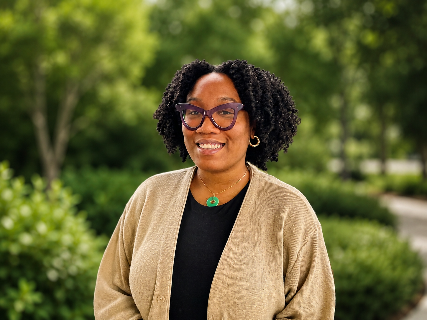 Smiling woman with curly hair and purple glasses wearing a tan cardigan outdoors among green trees.