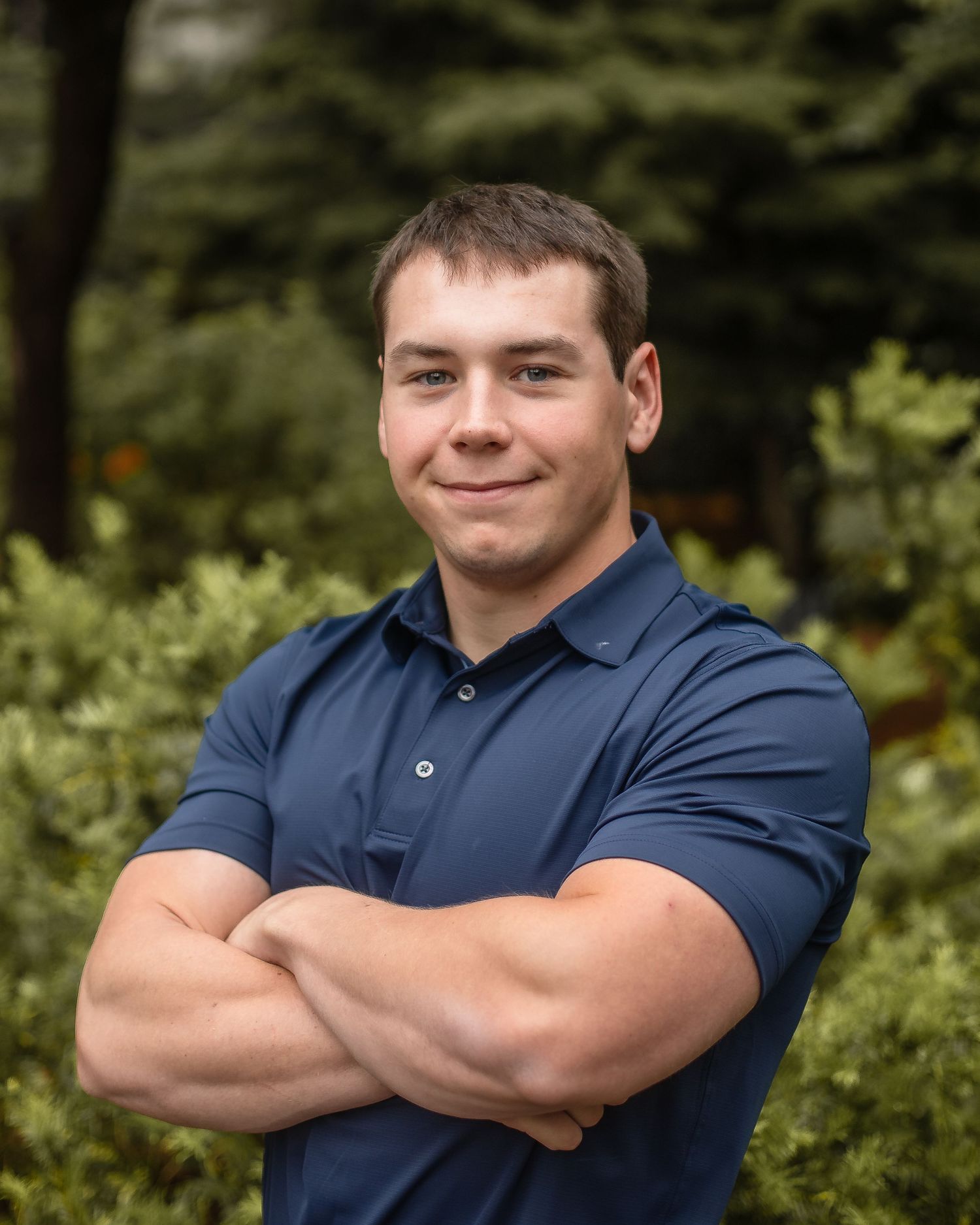 Young man in navy polo shirt stands confidently with arms crossed, smiling outdoors with green foliage background.