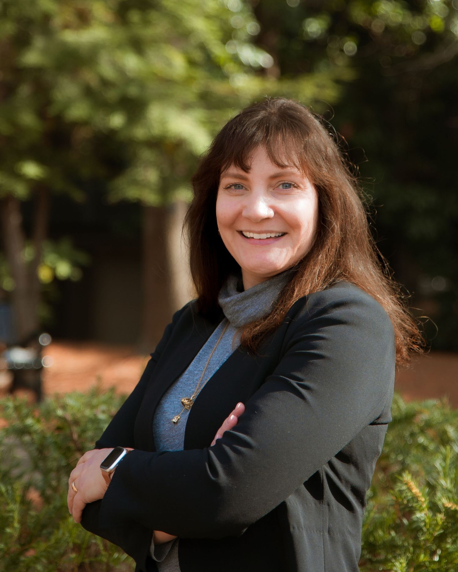 Professional outdoor portrait of someone in a black blazer and blue top smiling warmly against a natural green background.