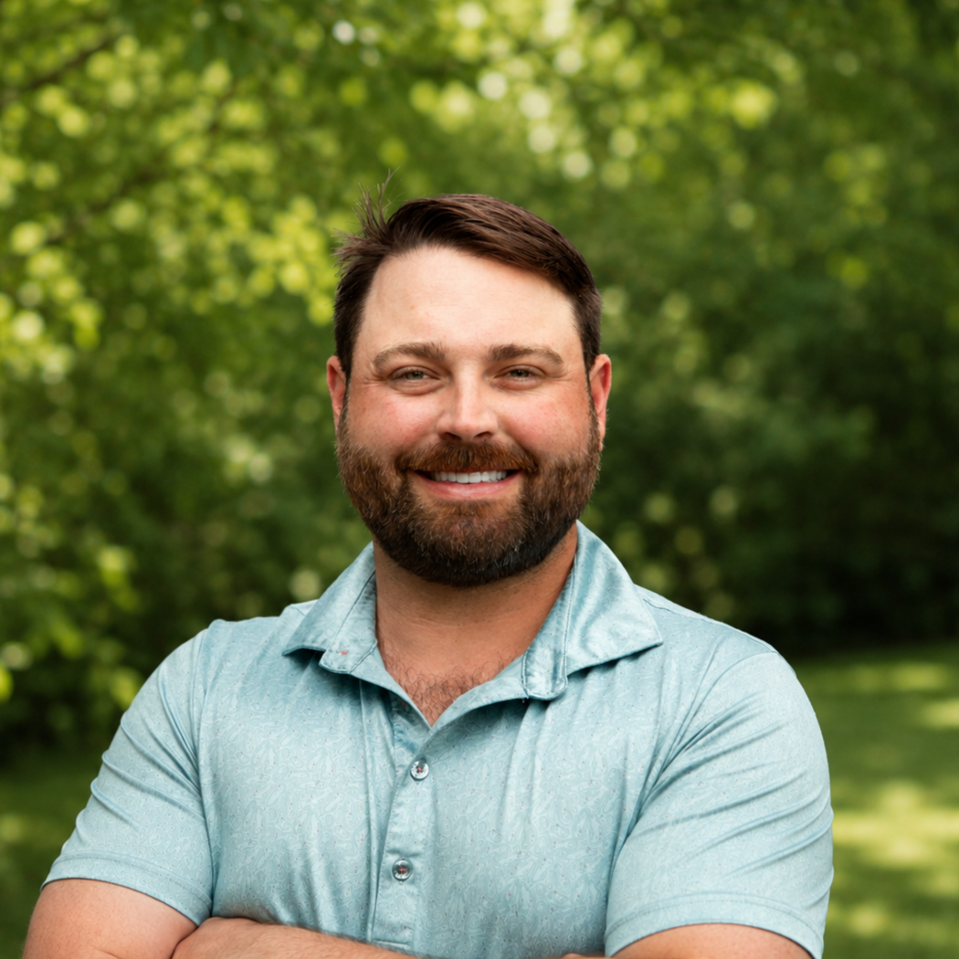 Smiling man with beard wearing light blue polo shirt standing outdoors with green trees in background.