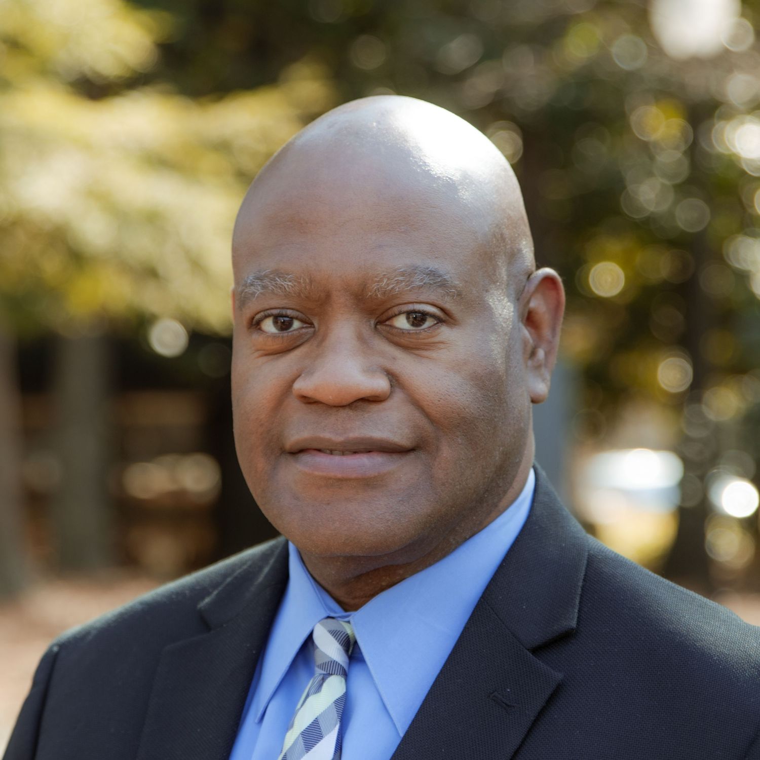 A person in a dark business suit smiles warmly against a blurred outdoor background with trees.