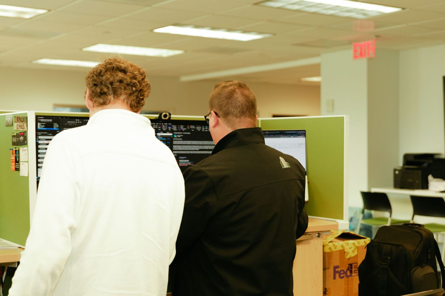 Office workers sit at computers in an open plan workspace with fluorescent lighting and white desks.