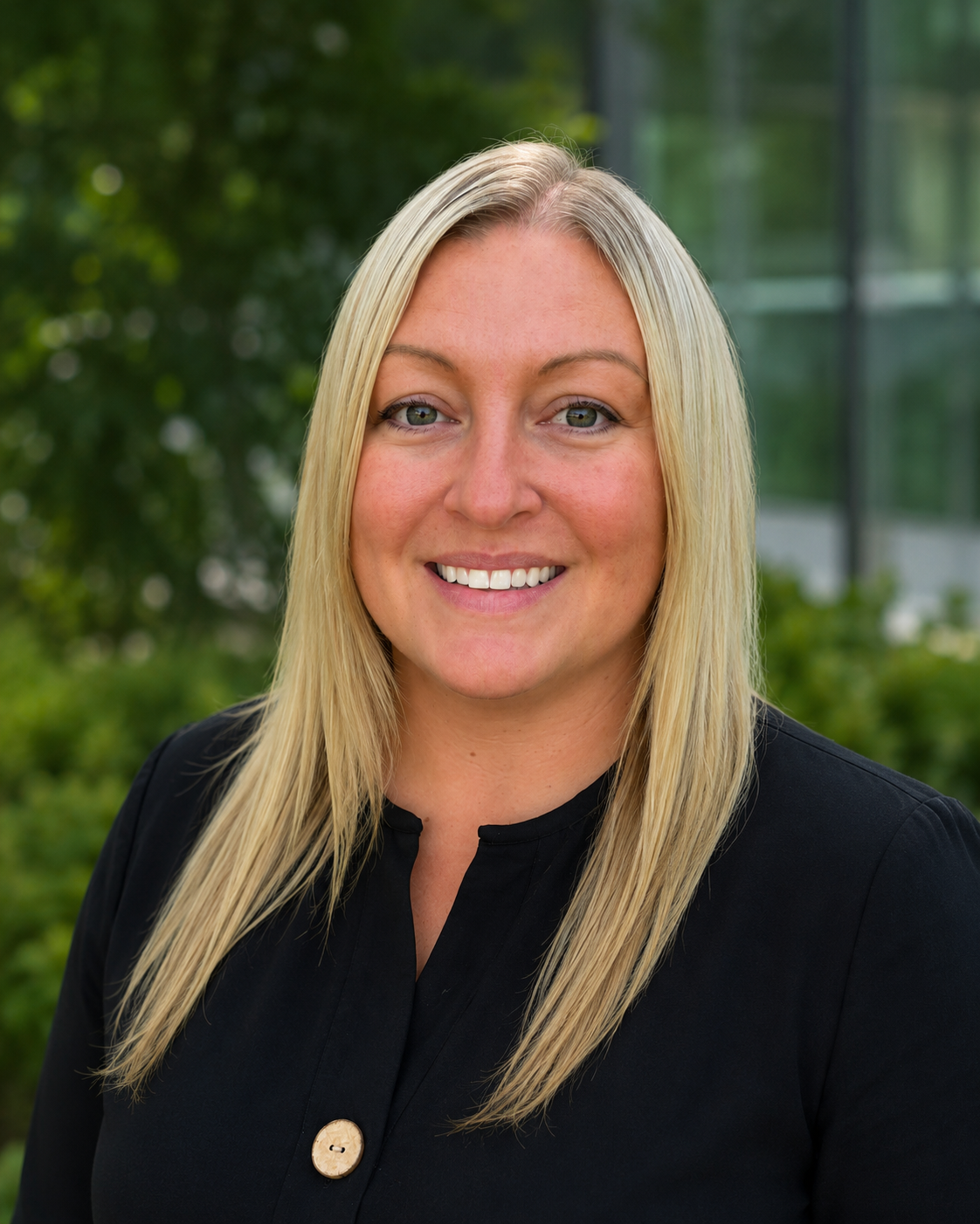 Professional headshot of a smiling blonde woman in a black blazer outdoors with green foliage background.