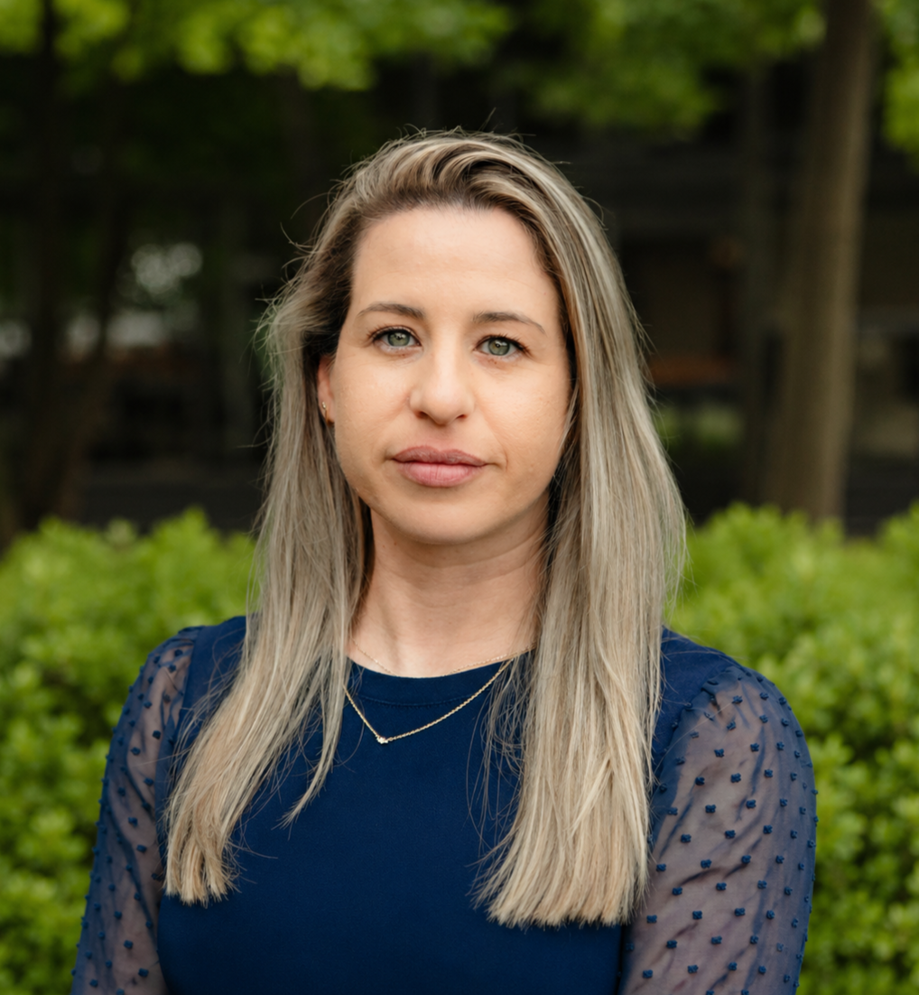 Professional headshot of a blonde woman in a navy blue top, posing outdoors with green foliage background.