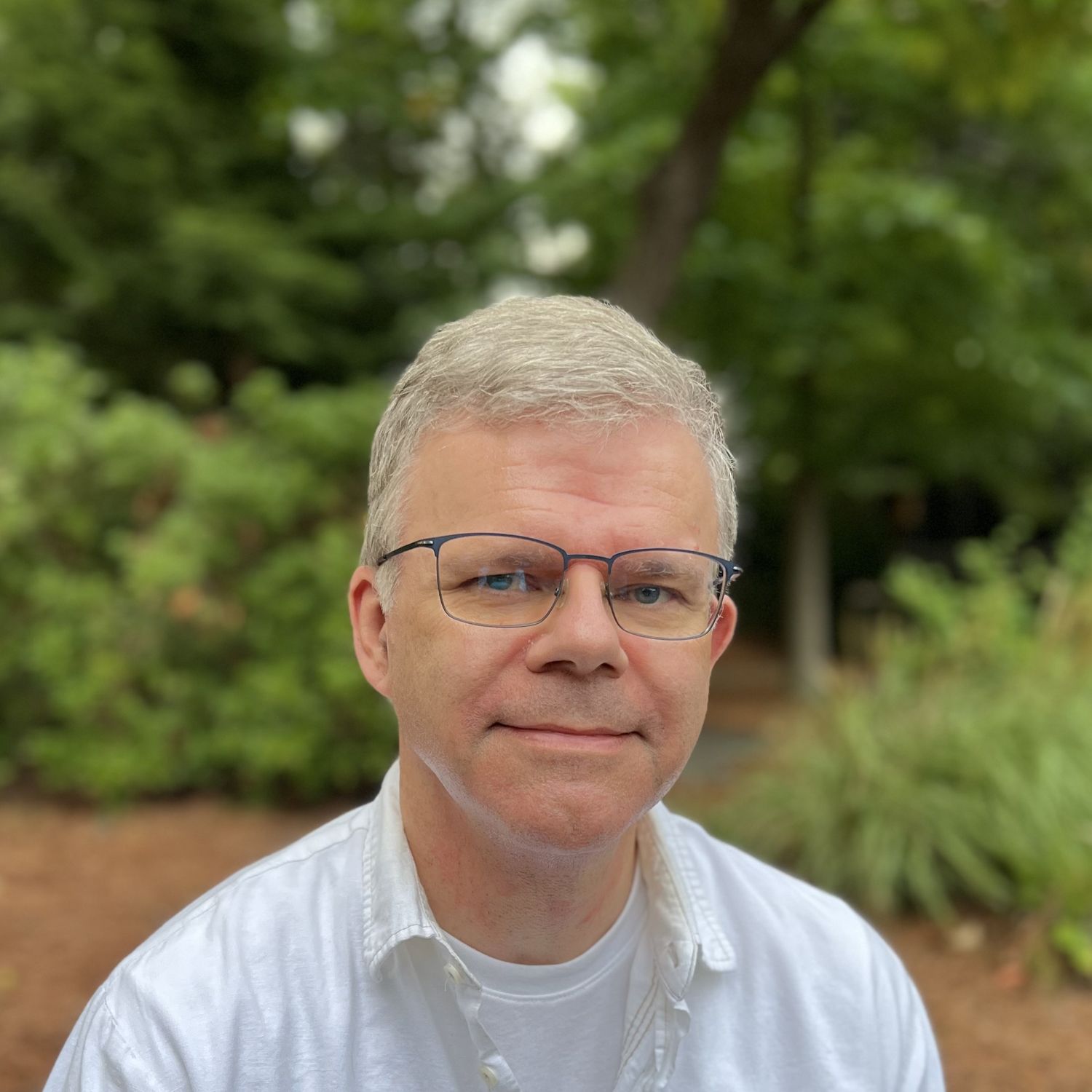 Person in white shirt smiling outdoors with trees and greenery in background.