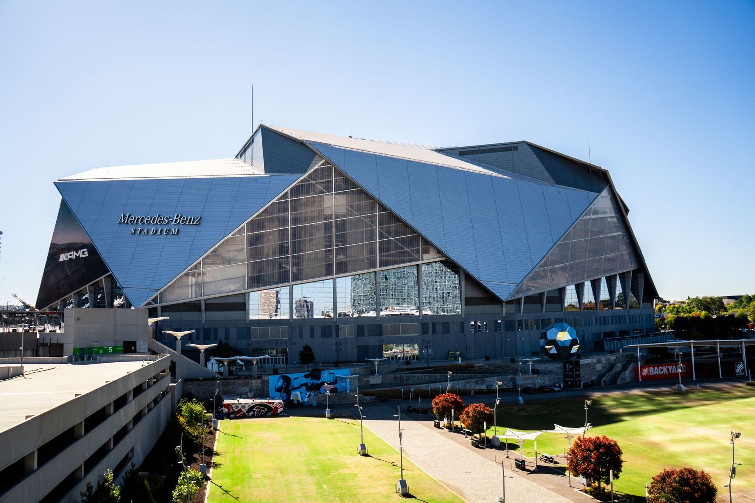 Modern angular stadium architecture with glass panels and geometric shapes against a bright blue sky.