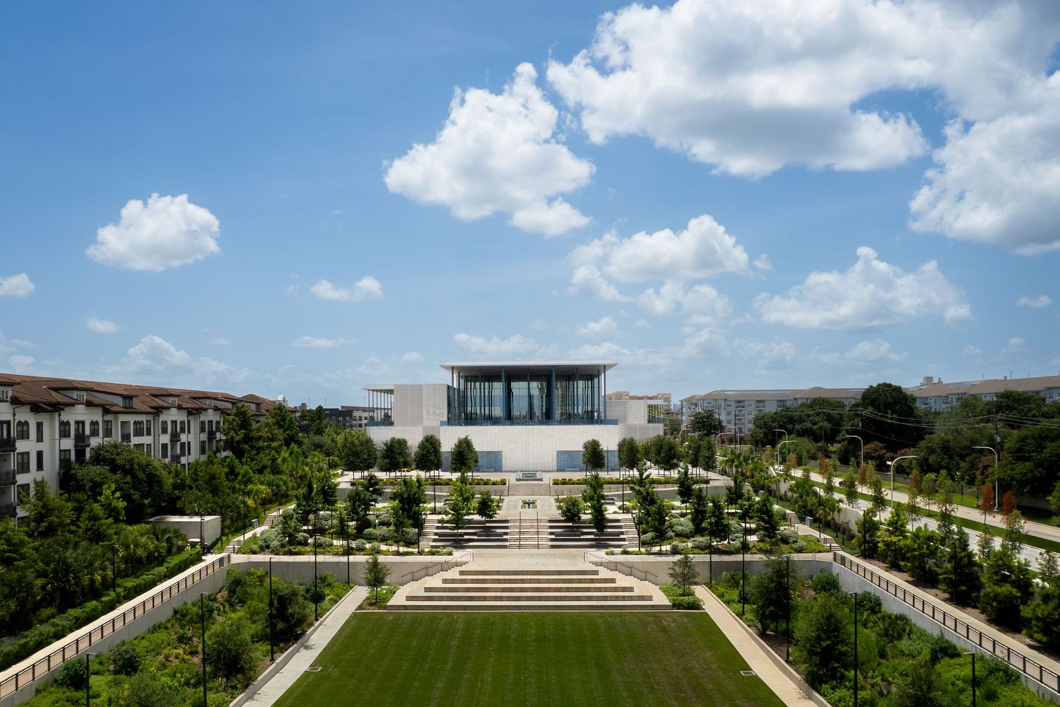 Aerial view of a modern white pavilion surrounded by manicured gardens, green lawns, and tree-lined pathways under a blue sky.
