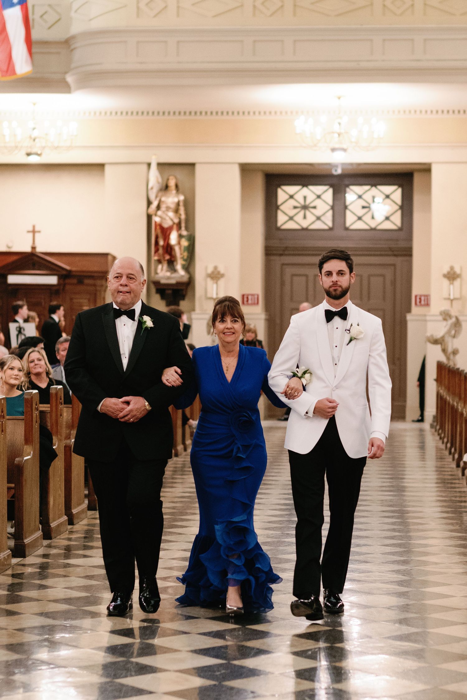 Wedding party members walk down a grand church aisle with checkered floors.