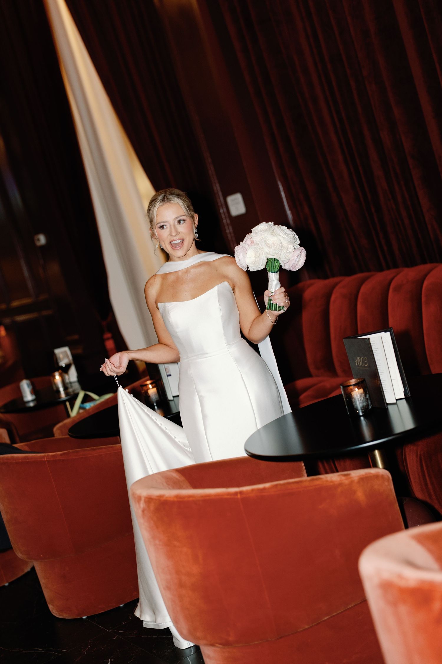 Bride in sleek white gown poses in red velvet booth at reception venue.