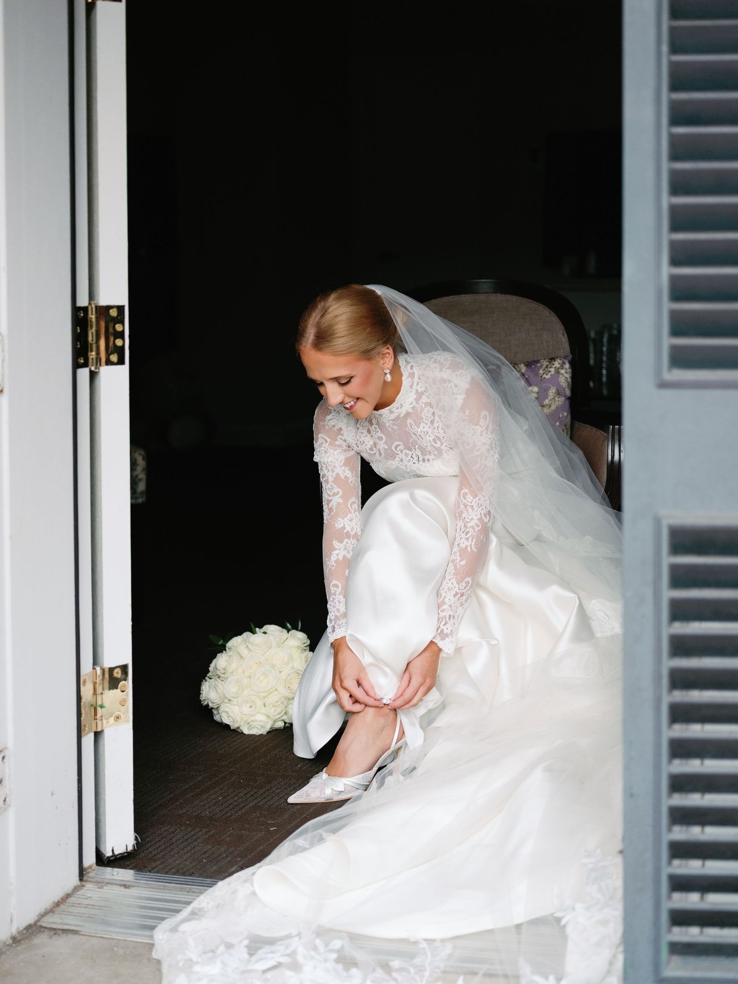Bride in elegant lace wedding gown adjusts her shoe while sitting in a doorway.