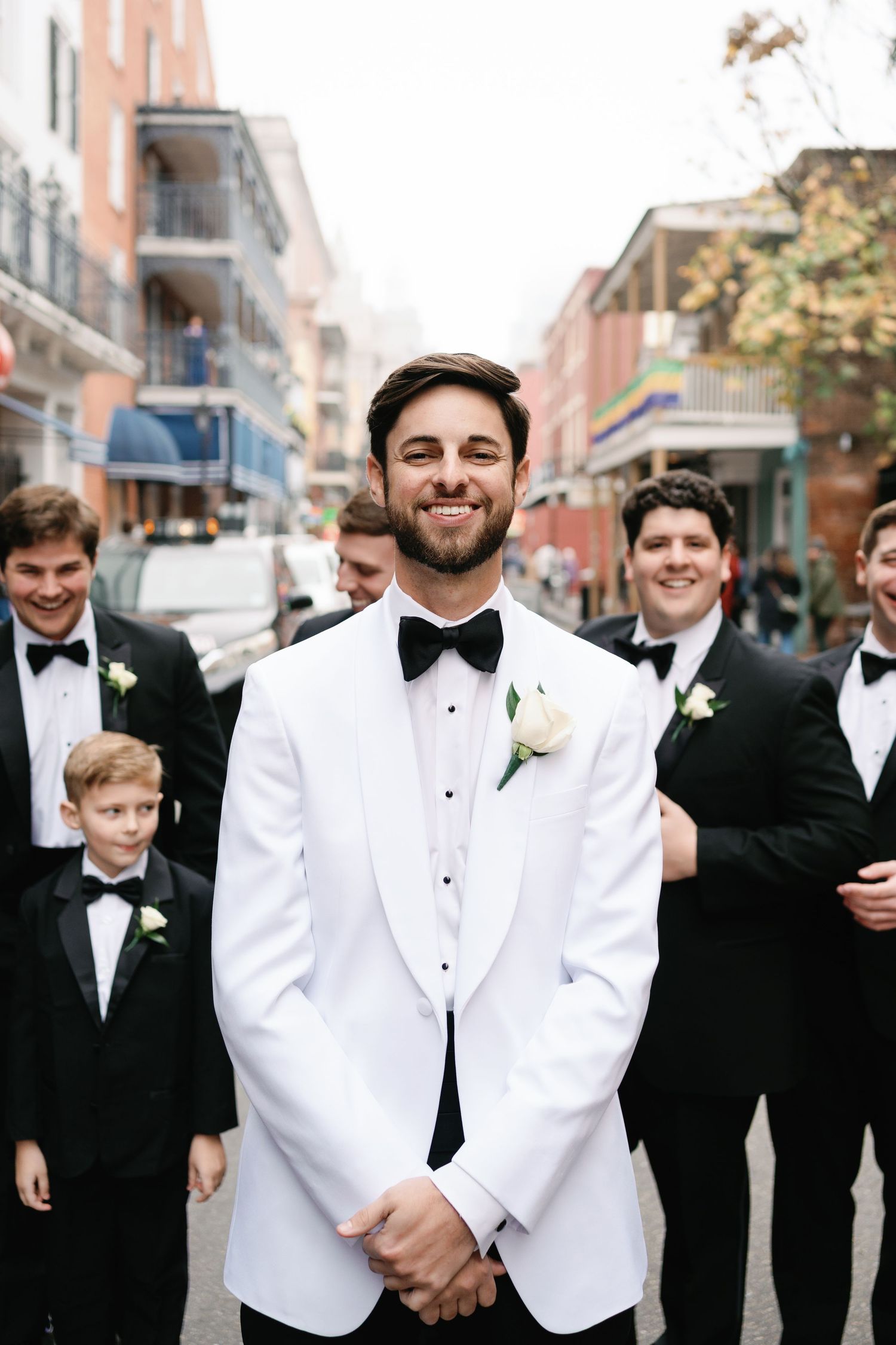 Groom in white tuxedo jacket stands with groomsmen on city street.