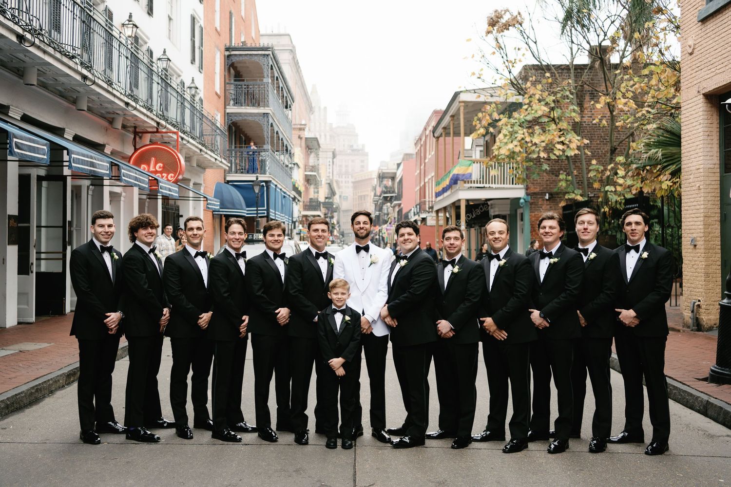 Large wedding party in black tuxedos posed along city street.