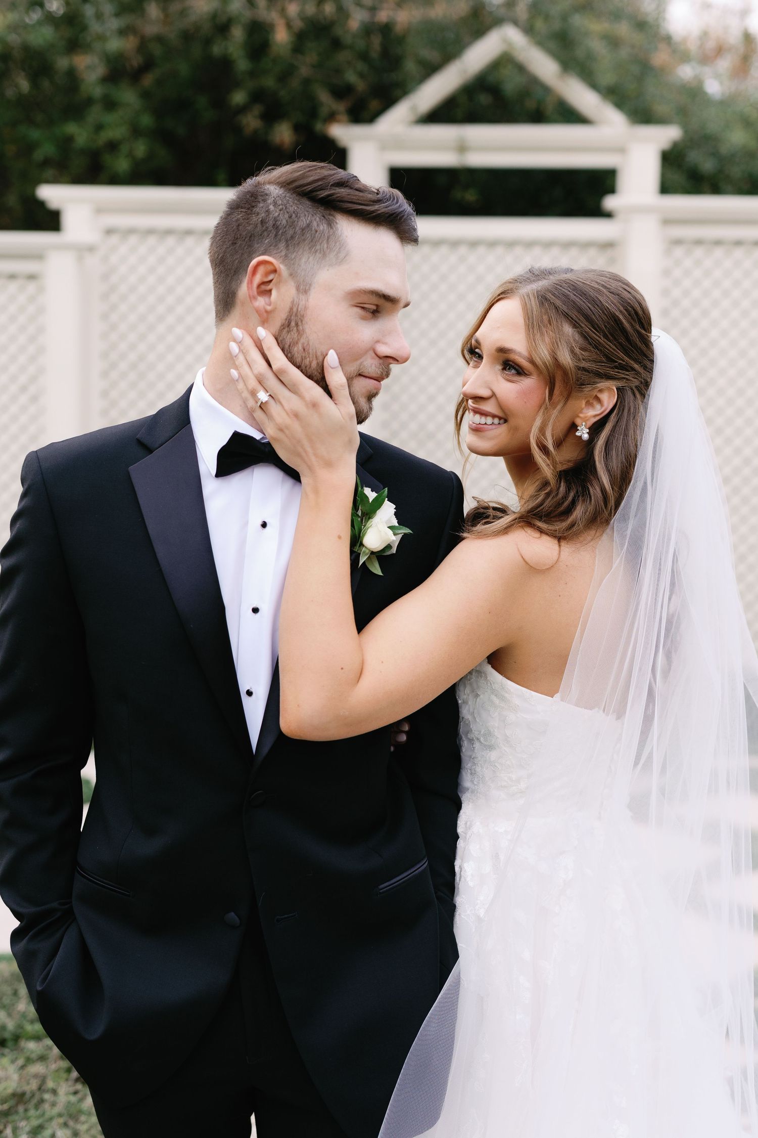A romantic wedding couple shares intimate moments in front of a white lattice fence outdoors.