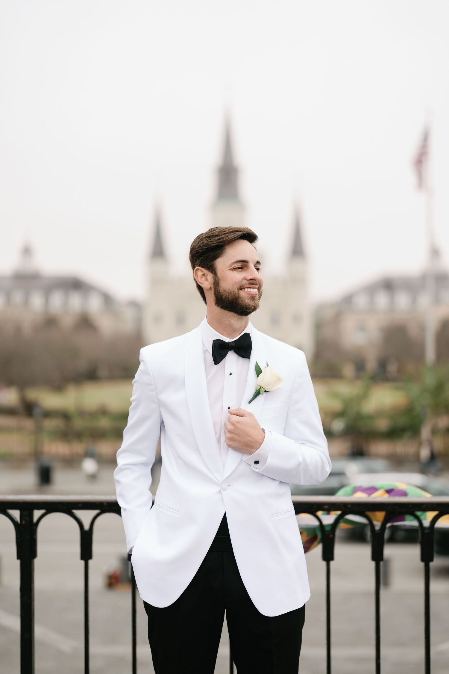 A groom in a white tuxedo jacket and black bow tie smiles while standing on a balcony with a church spire in the background.