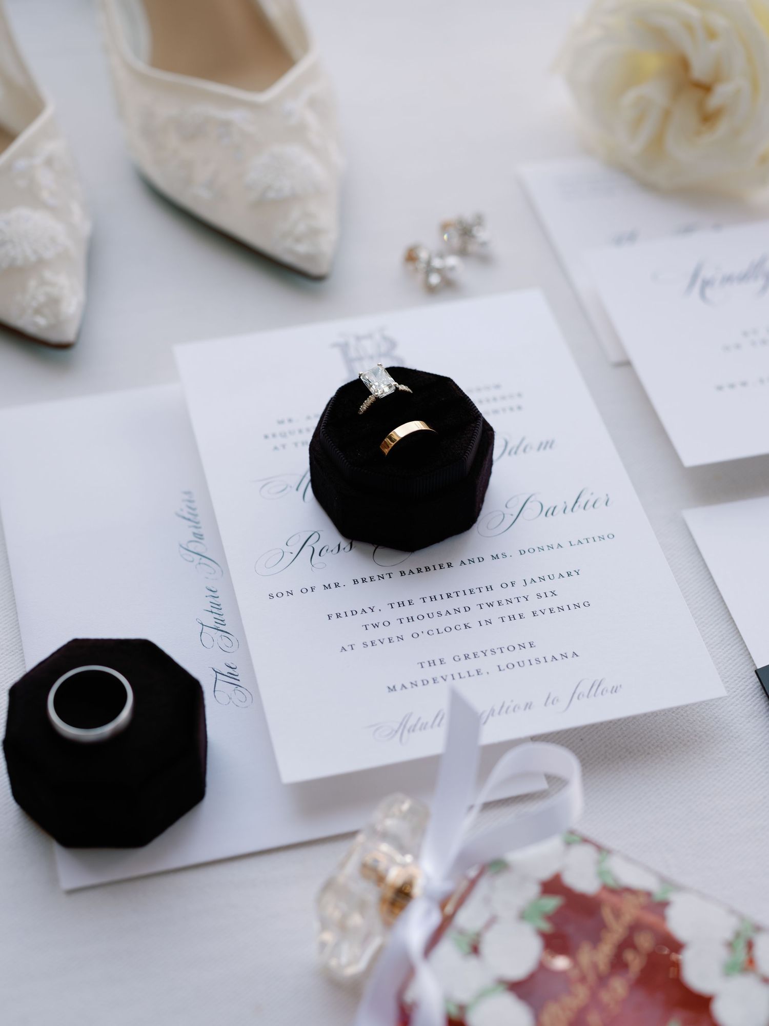 Elegant white and black wedding details including lace shoes, invitations, and ring boxes on a light background.