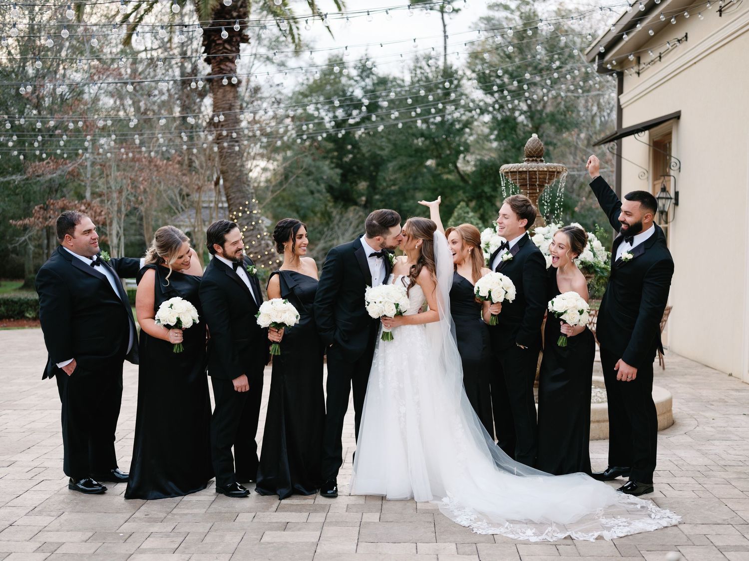 Wedding party in black attire poses together on stone patio.