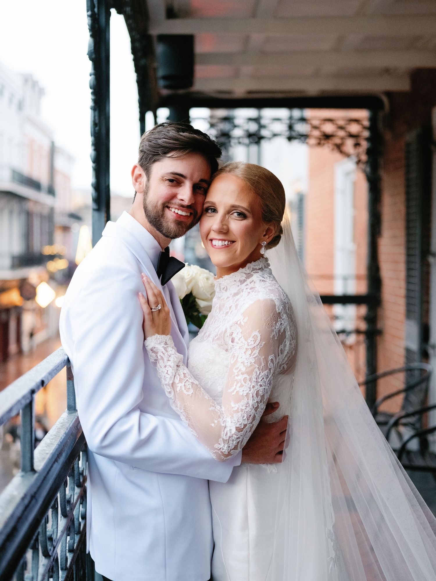 Newlyweds embrace lovingly while wearing white formal attire on a vintage-style porch.