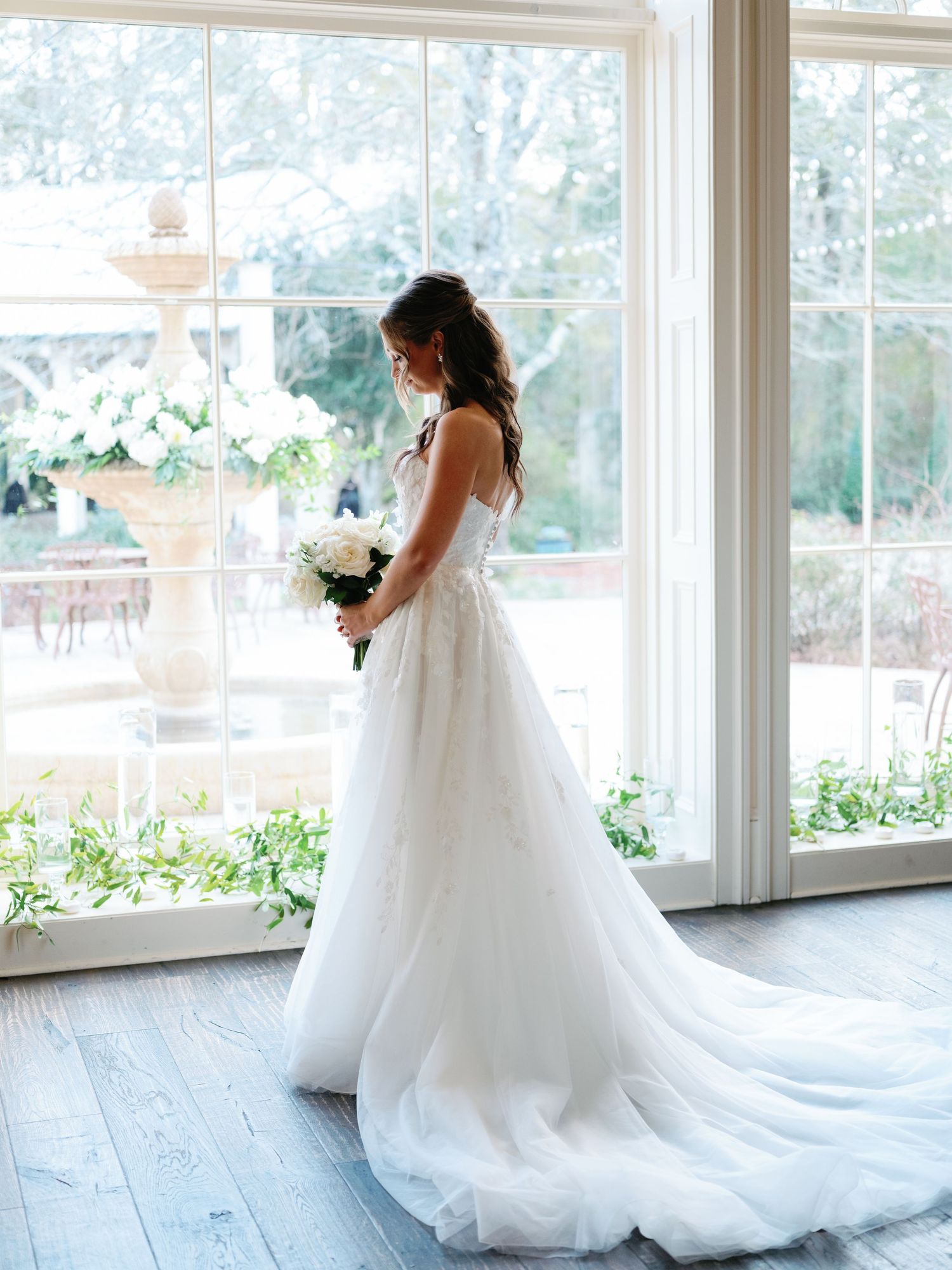 A bride in a flowing white wedding gown stands by large windows holding a white bouquet.