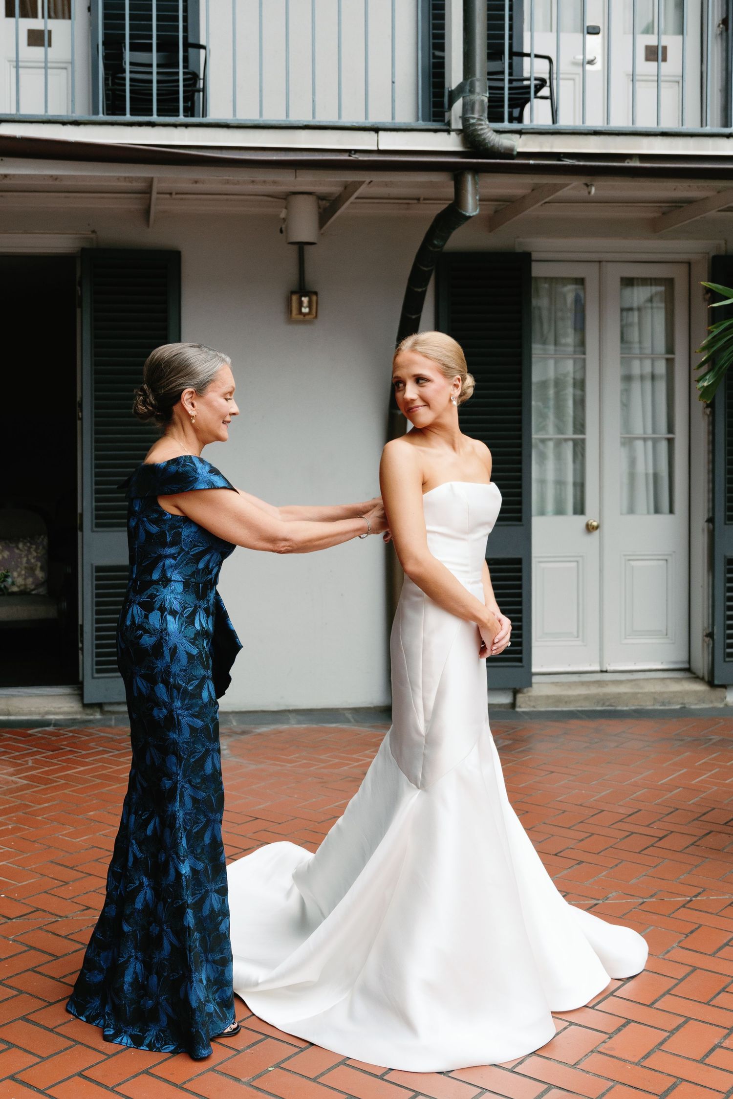 A woman in navy blue helps adjust a white wedding dress on a brick patio.