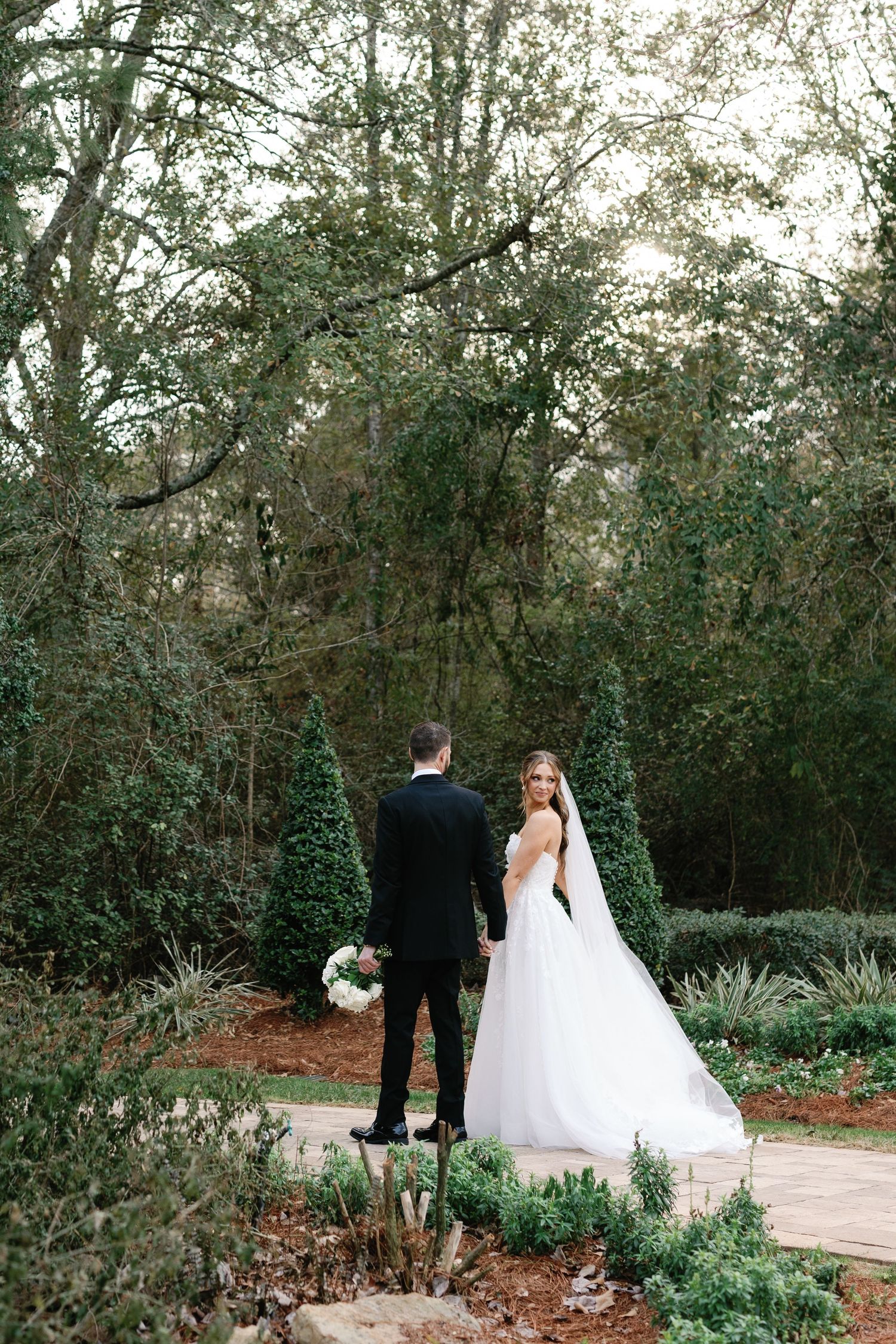 A couple stands on a garden path surrounded by lush greenery on their wedding day.