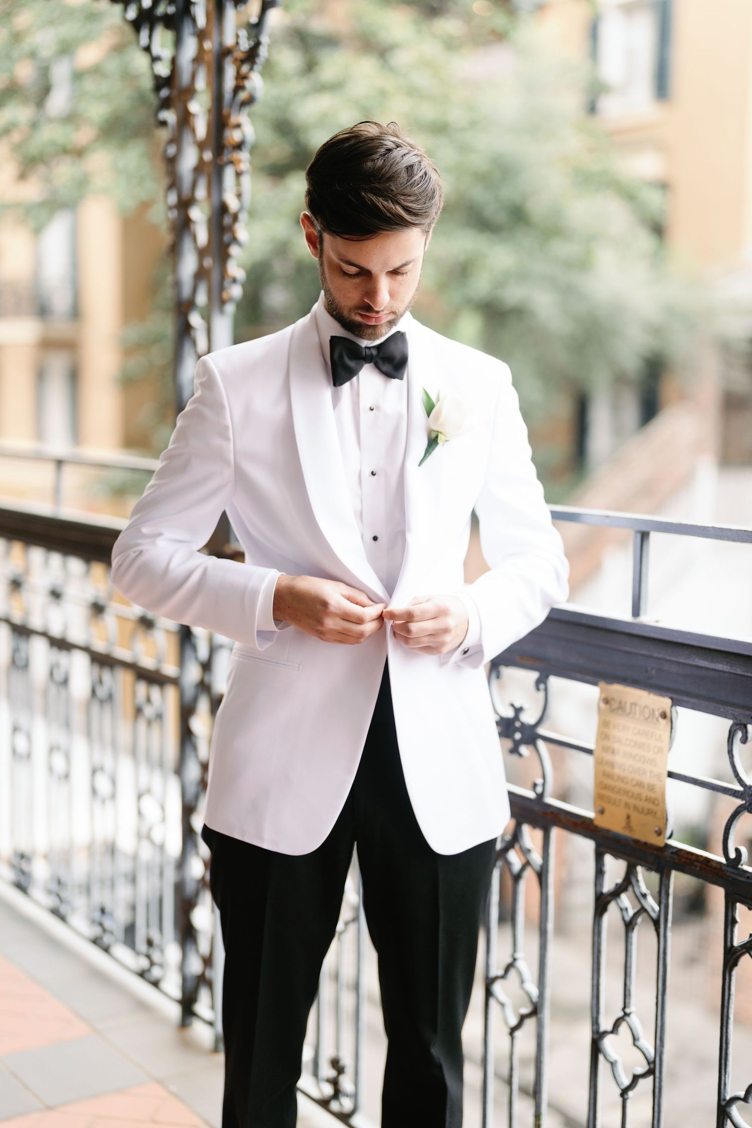 Groom adjusts his attire while standing on decorative balcony in white dinner jacket.