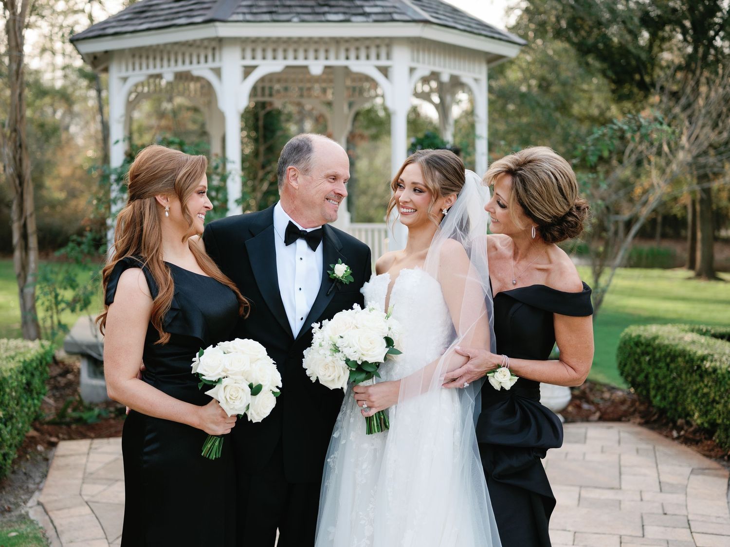 Wedding party members in formal attire gather outside a white gazebo for photos.