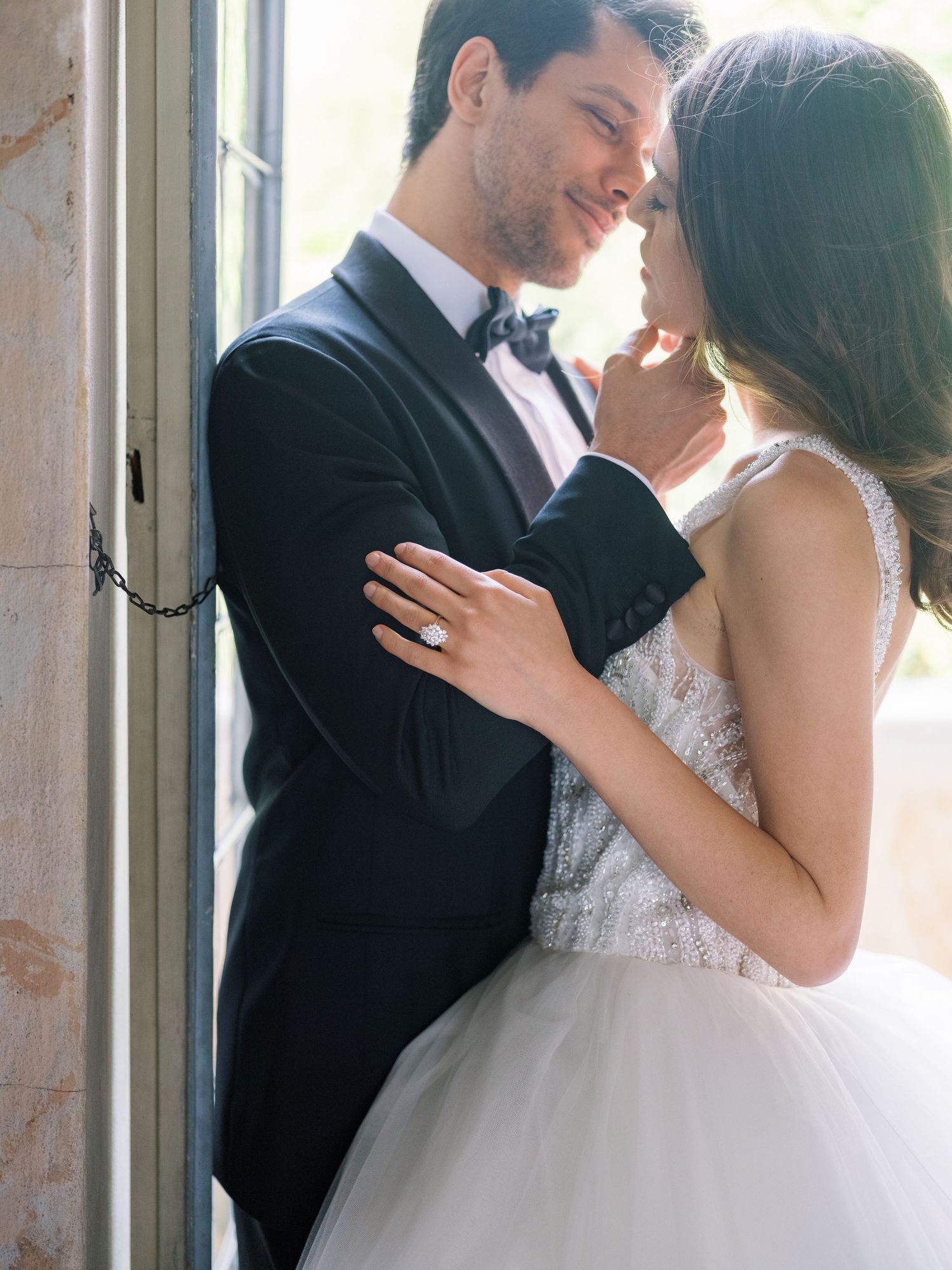 Elegant couple shares intimate moment in formal wedding attire by sunlit window.