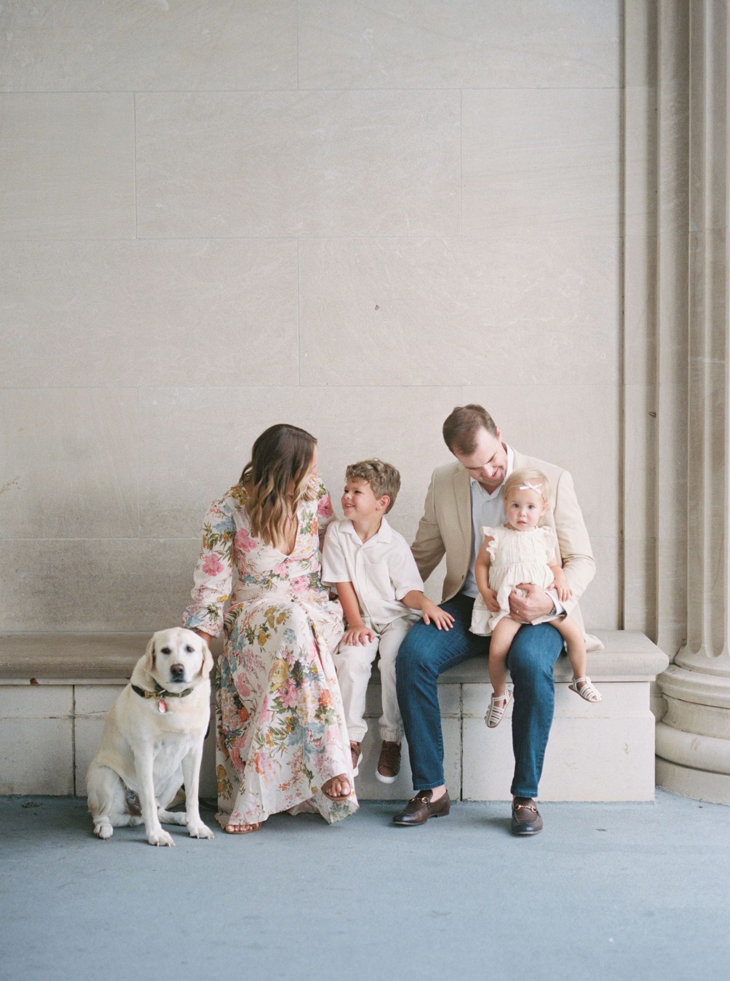Family sits together on stone ledge with their yellow Labrador retriever dog in elegant outdoor portrait setting.