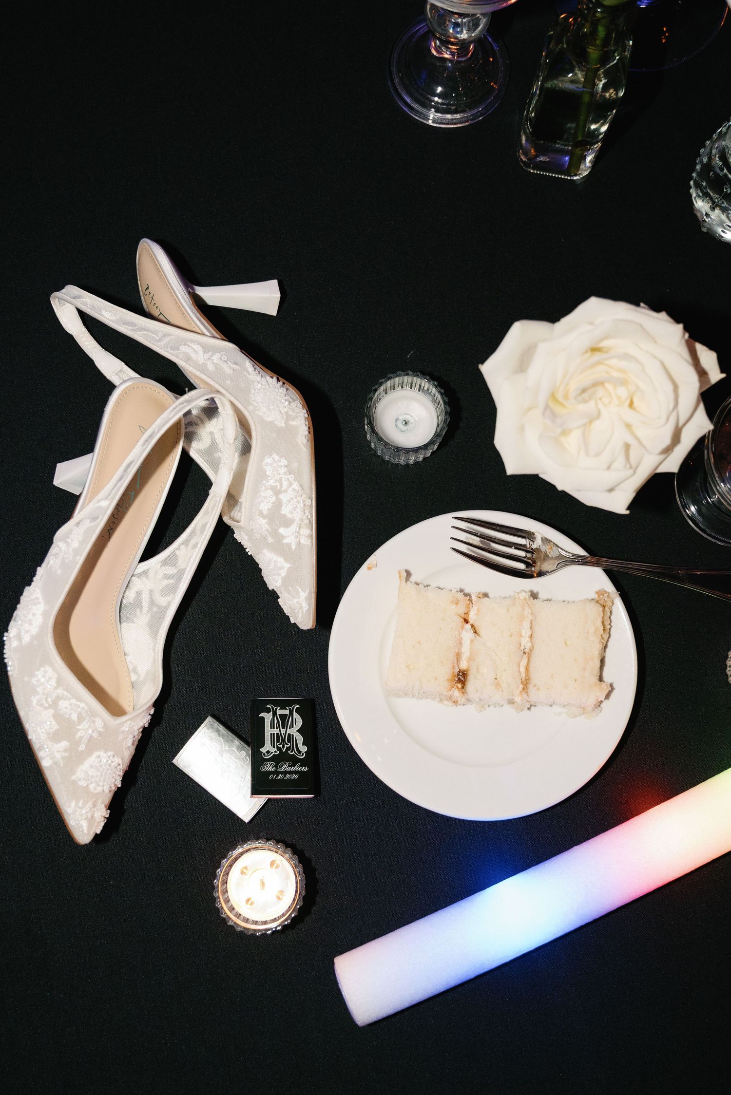 Wedding details flatlay with lace heels, cake slice, and white rose on black background.