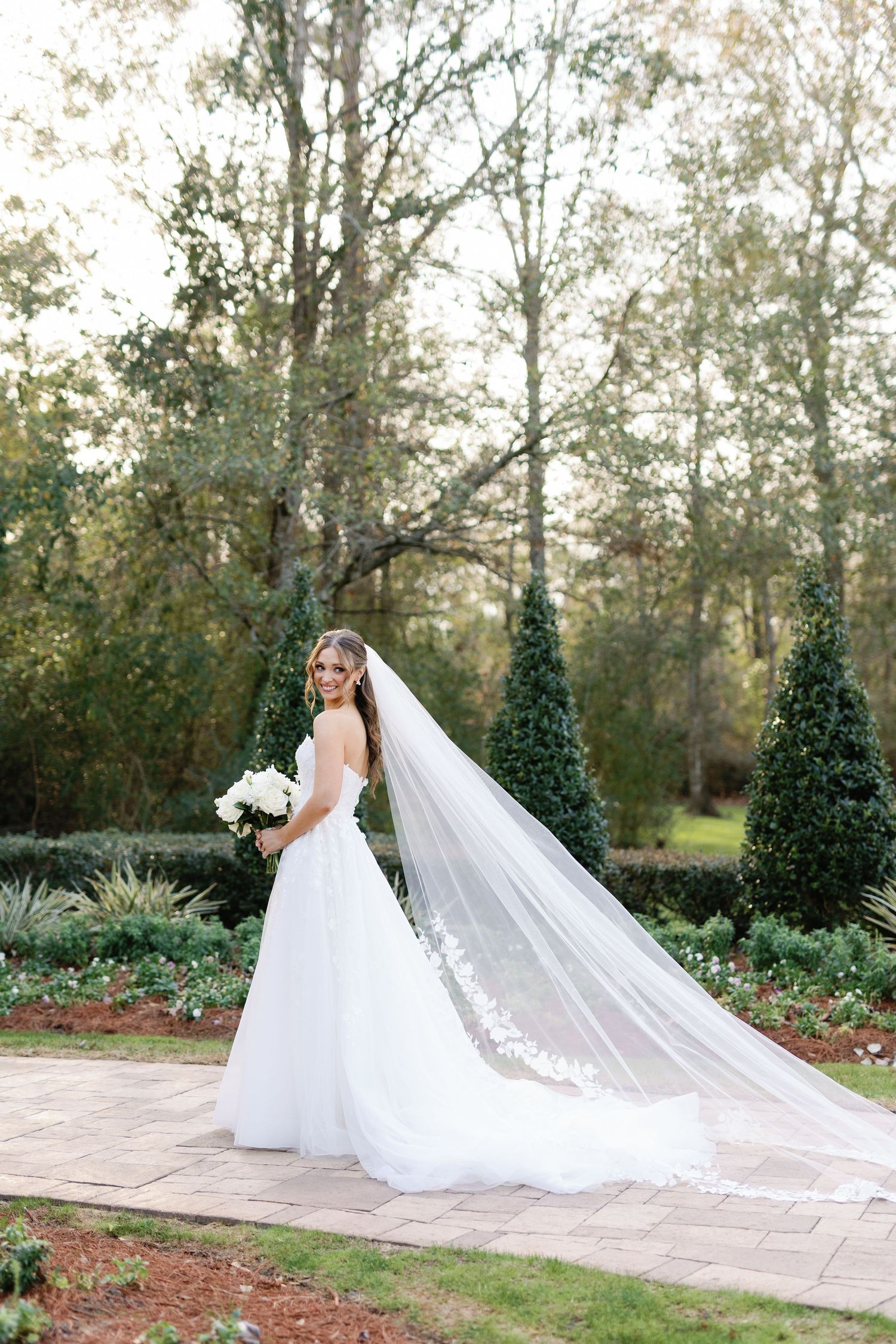 A bride in a flowing white wedding gown with a dramatic cathedral-length veil poses in a garden setting with topiary trees.
