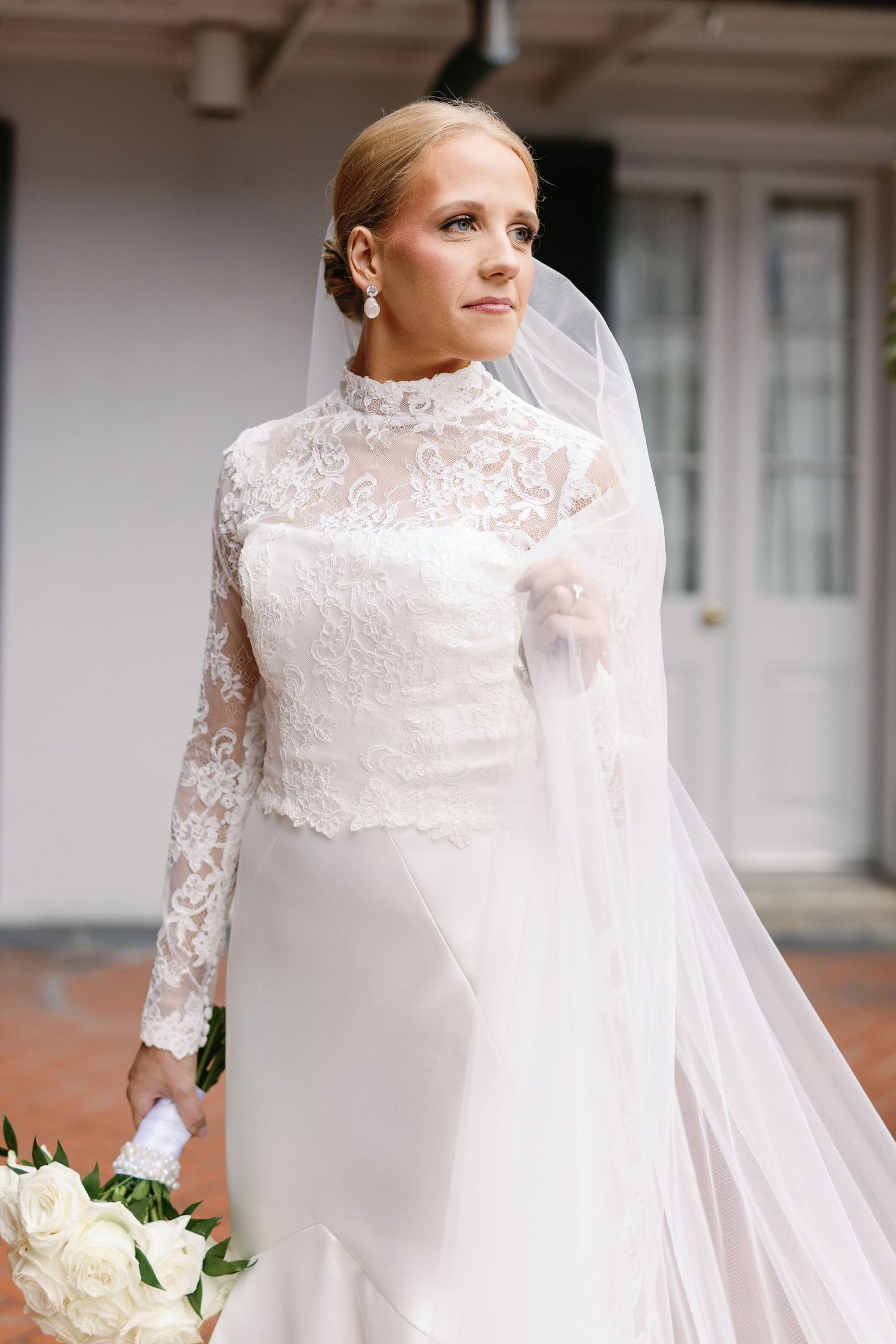 Elegant bride in long-sleeved lace wedding gown with flowing veil holds white bouquet on brick porch.