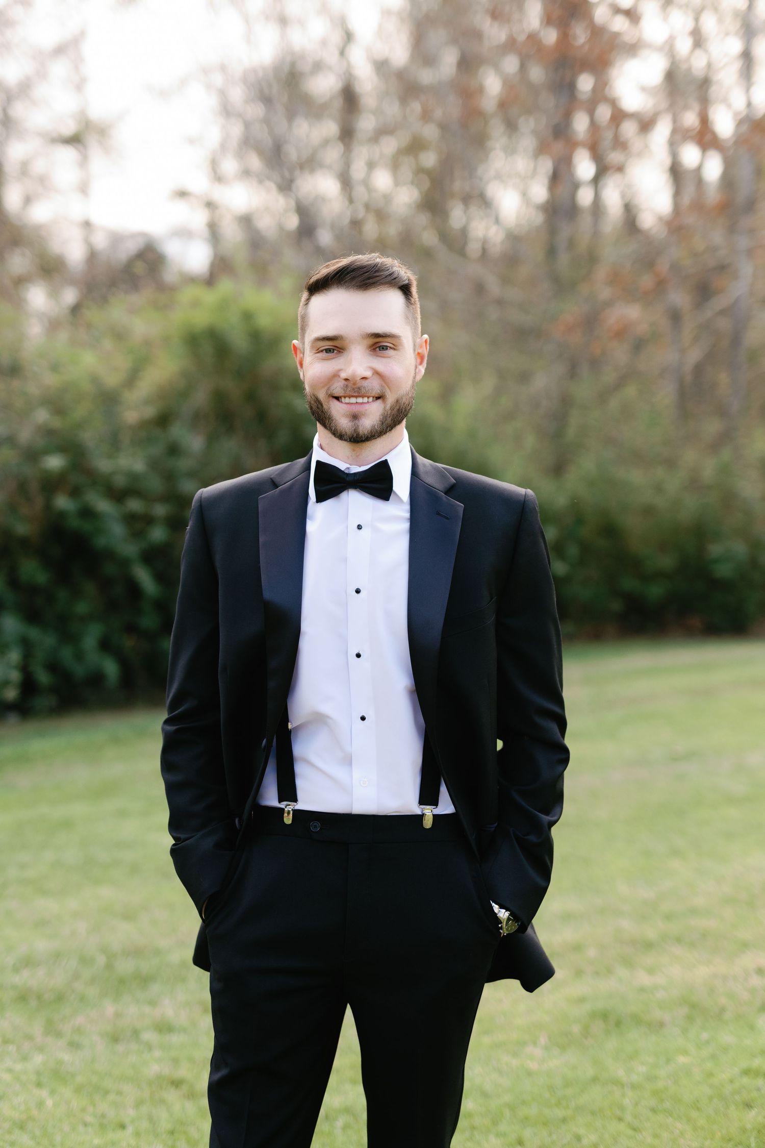 A groom in a classic black tuxedo and bow tie stands on a lawn.