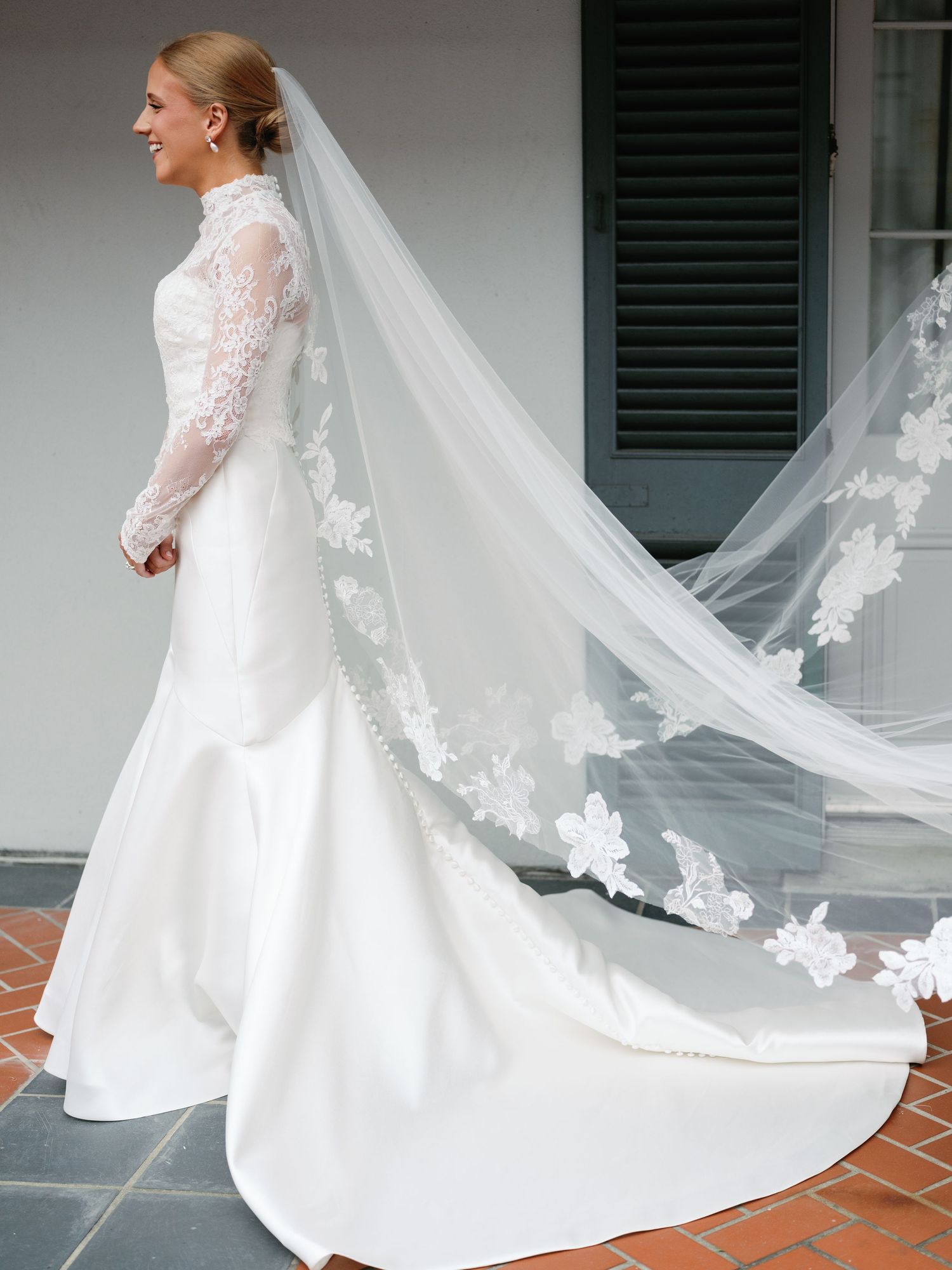 A flowing white wedding dress with lace details and long cathedral veil twirls elegantly on a porch.