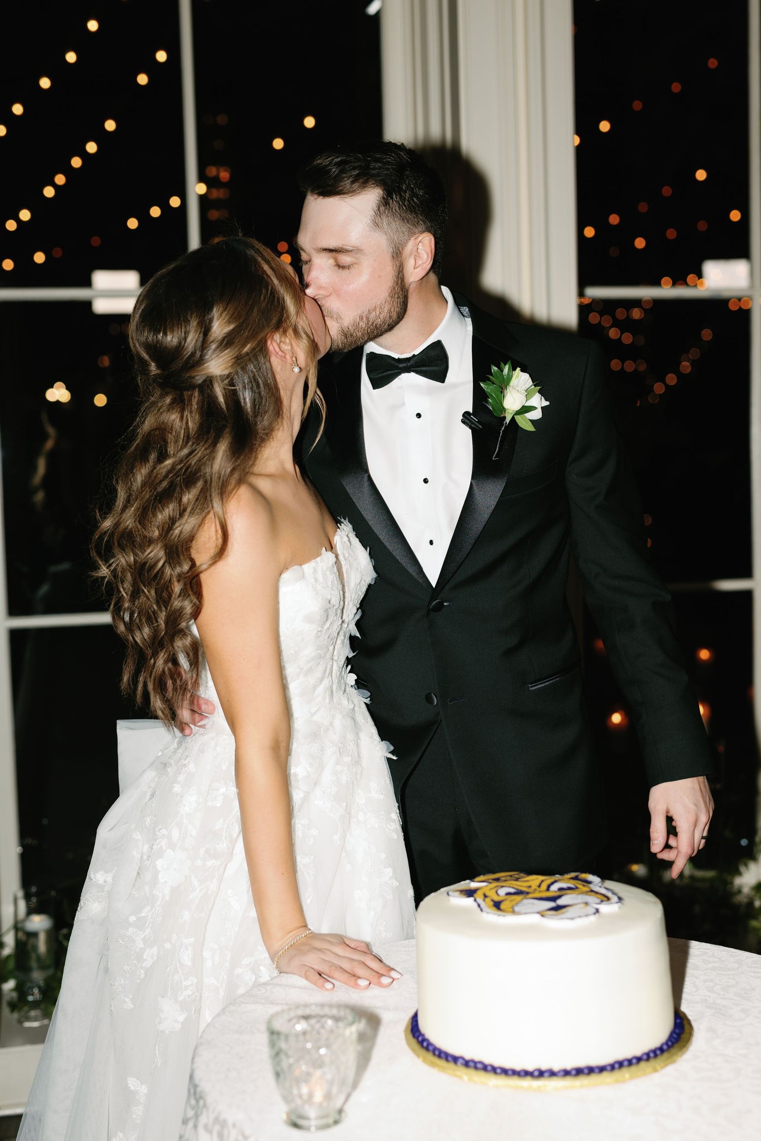 Newlywed couple in formal attire cuts their wedding cake together at an evening reception with twinkling lights.