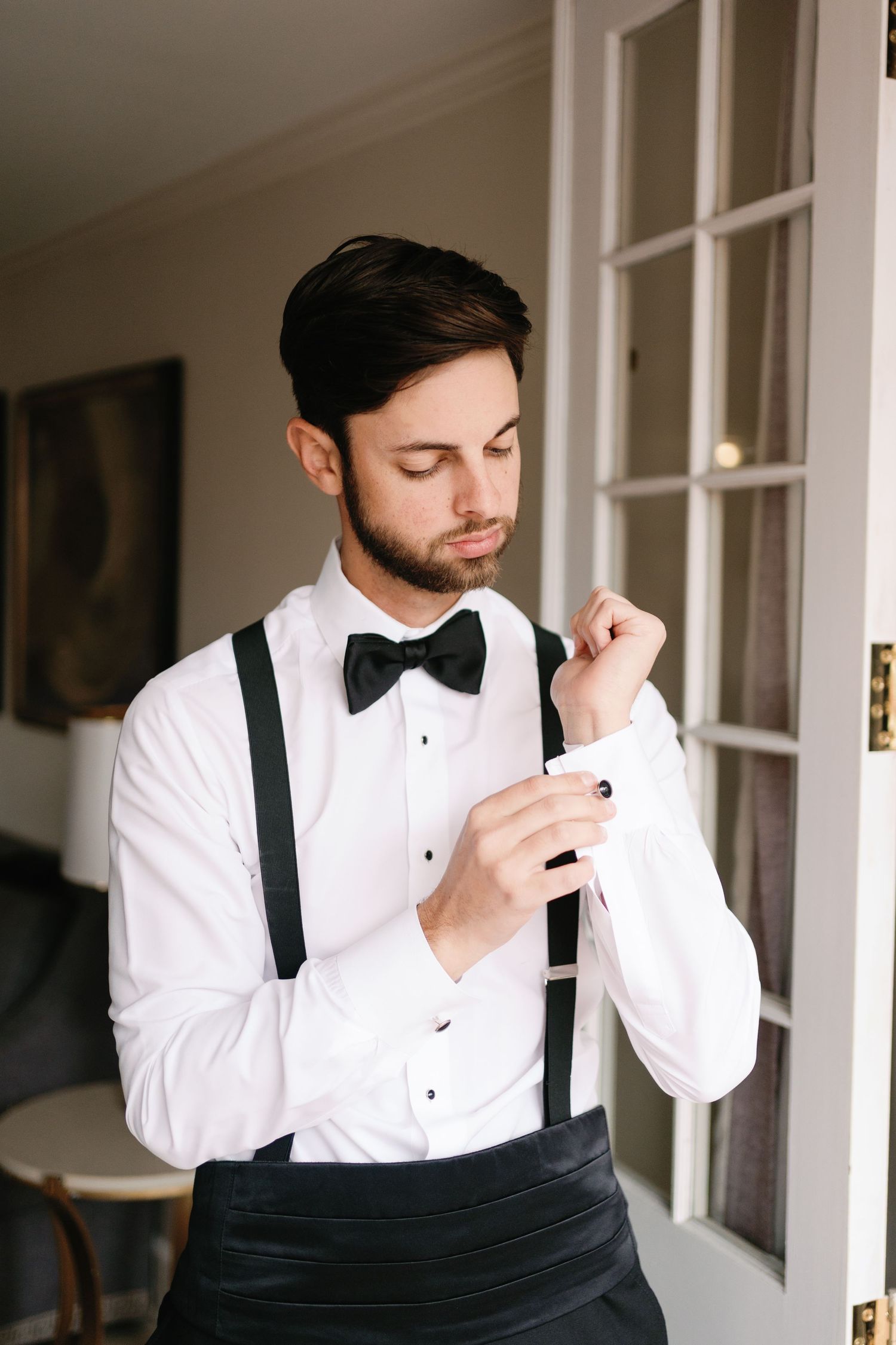 Groom in white tuxedo and suspenders preparing for wedding ceremony.