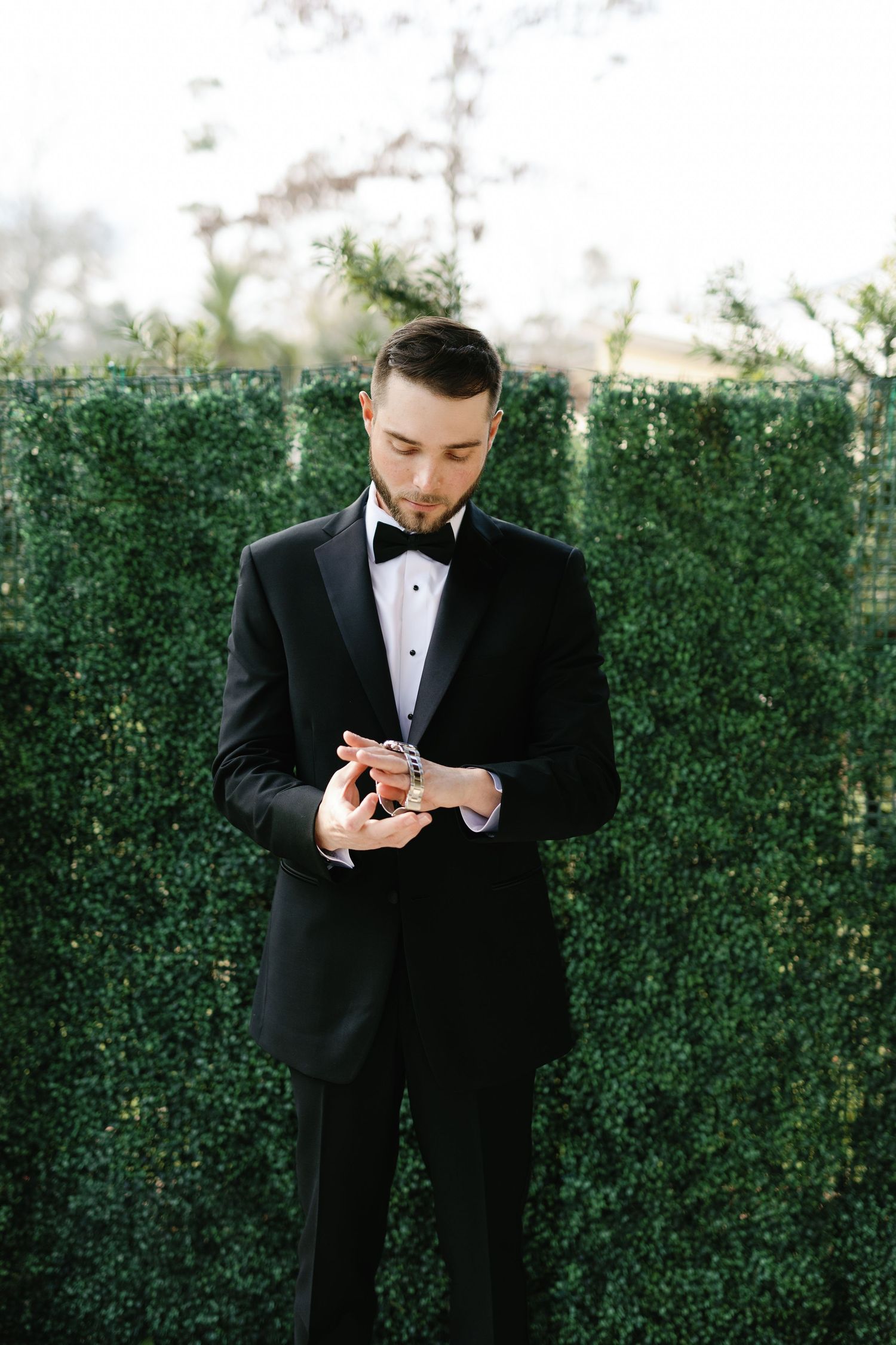 Groom in black tuxedo checking his phone while standing in front of a green hedge wall.