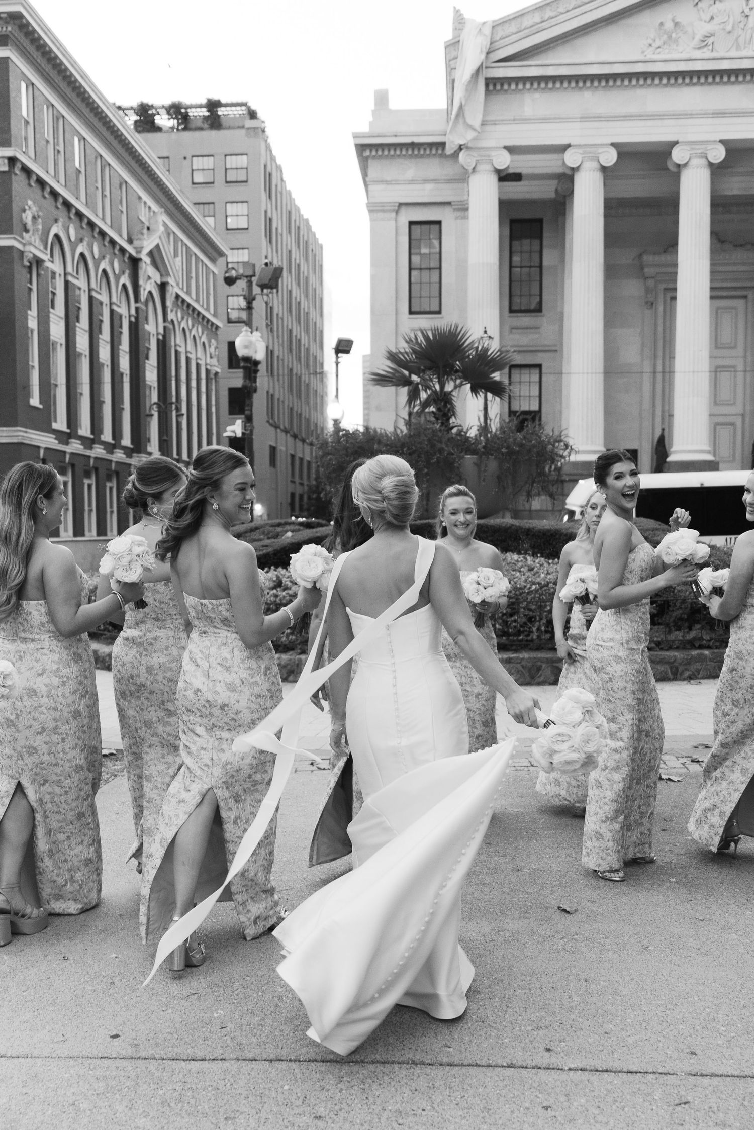 A group of bridesmaids in elegant dresses dance joyfully outside a classical building with columns.
