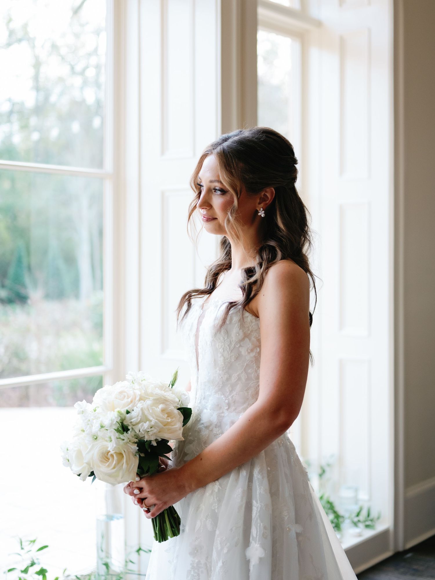 Bride in white lace wedding dress holds elegant white rose bouquet while standing by sunlit windows.