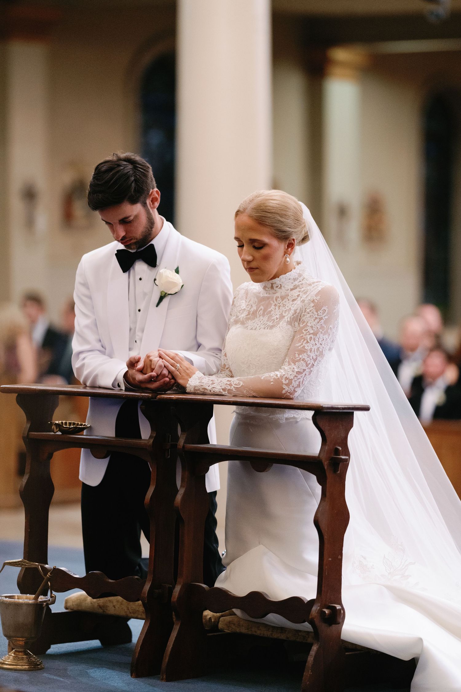 Bride and groom praying together at wooden church pews during ceremony.