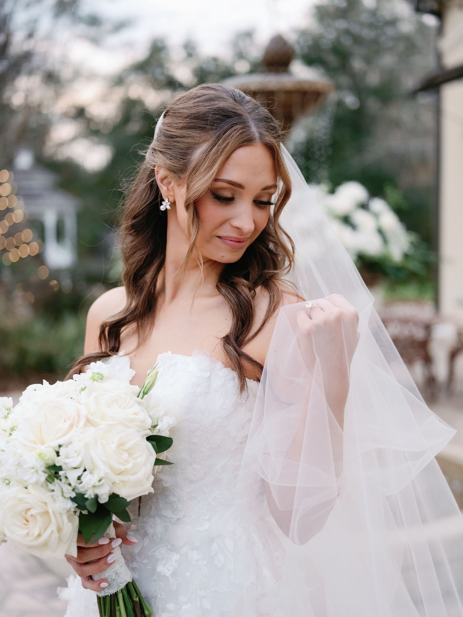 A bride in a white gown holds a bouquet of white roses while wearing a flowing veil in an outdoor setting.