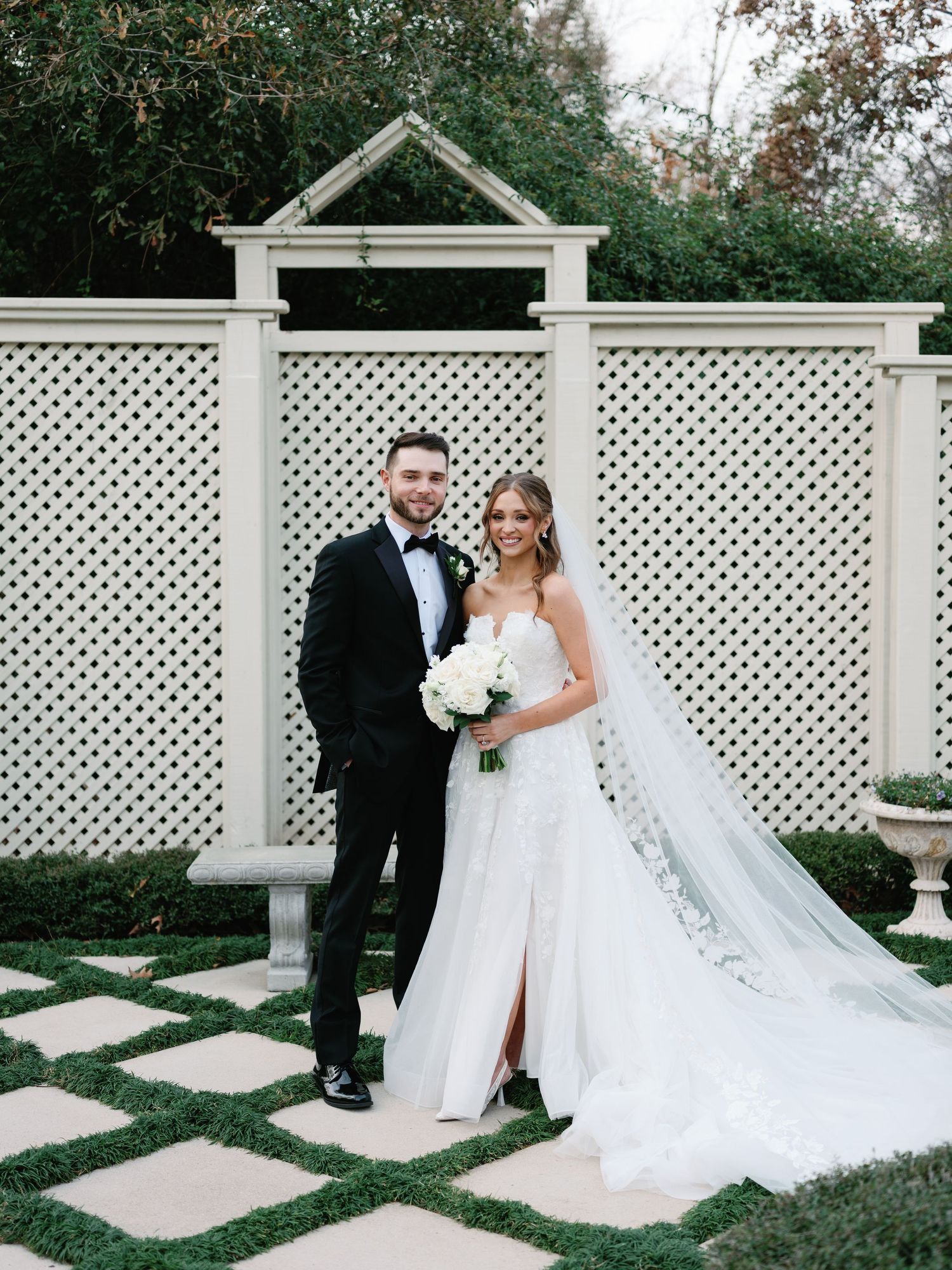 Elegant bride and groom pose for portraits along decorative white garden trellis walkway.