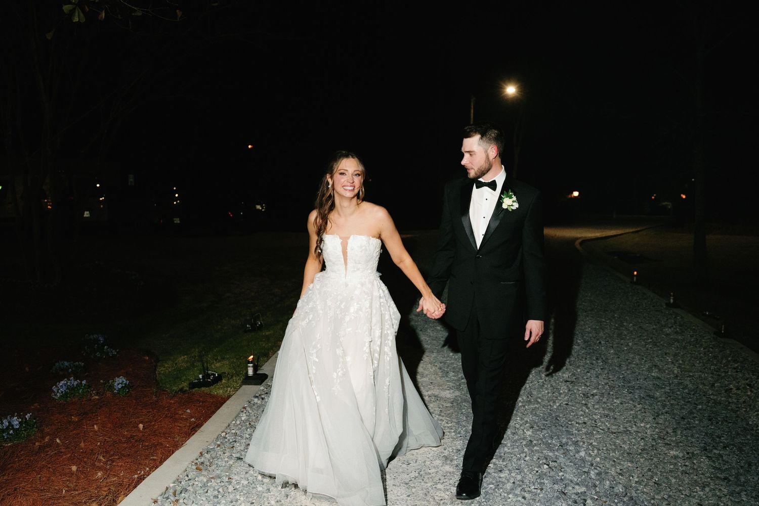 A couple walks hand in hand on a gravel path at night, the bride in a white gown and groom in a black tuxedo.