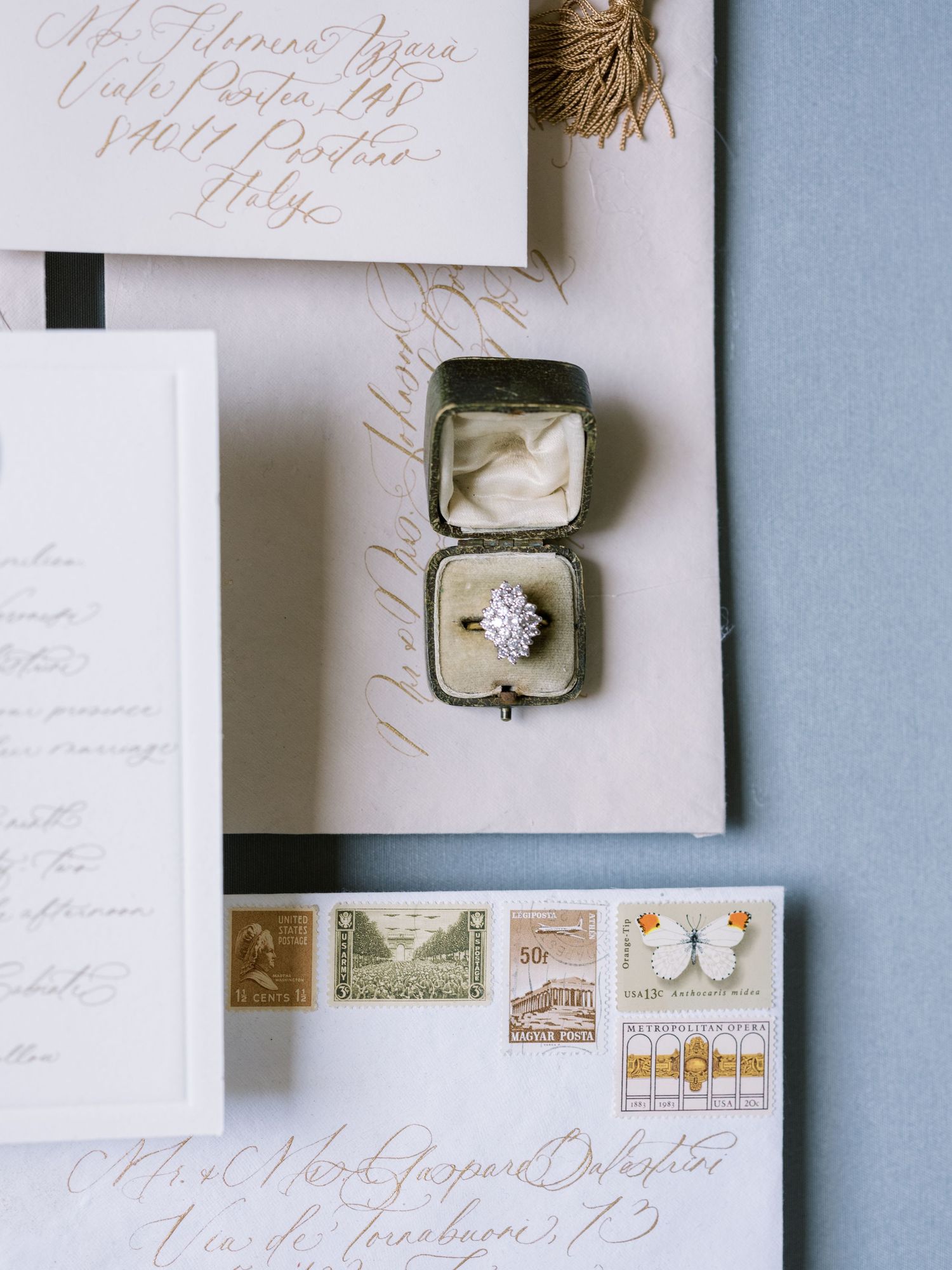 Wedding stationery flatlay with vintage stamps, ring box, and elegant calligraphy on light blue background.