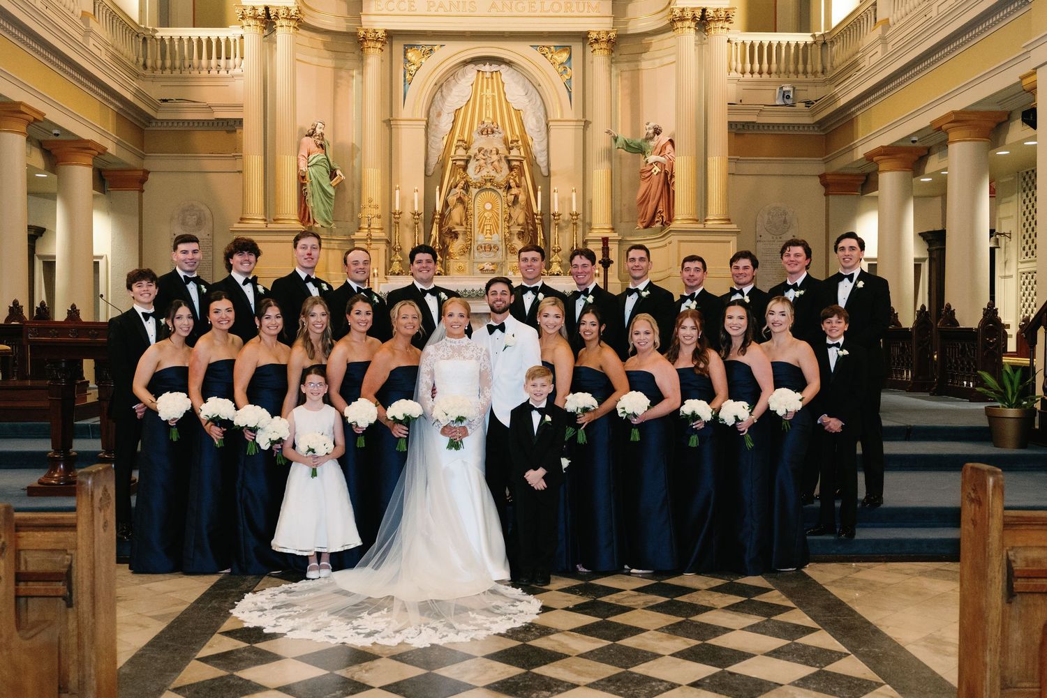 Large wedding party in black and white attire poses on church altar steps.