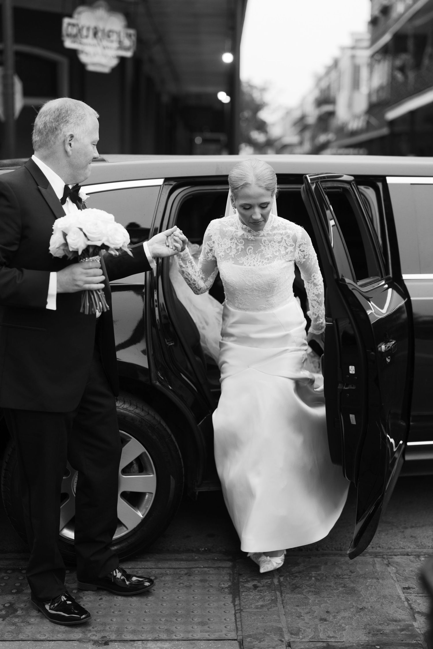 Black and white photo of bride in lace gown stepping out of vintage car with assistance.