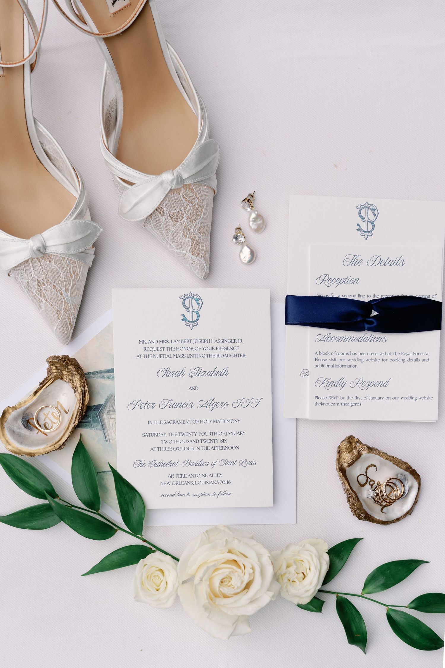 Elegant wedding invitation flatlay with white lace heels, navy ribbon details, oyster shell, and white roses with greenery.