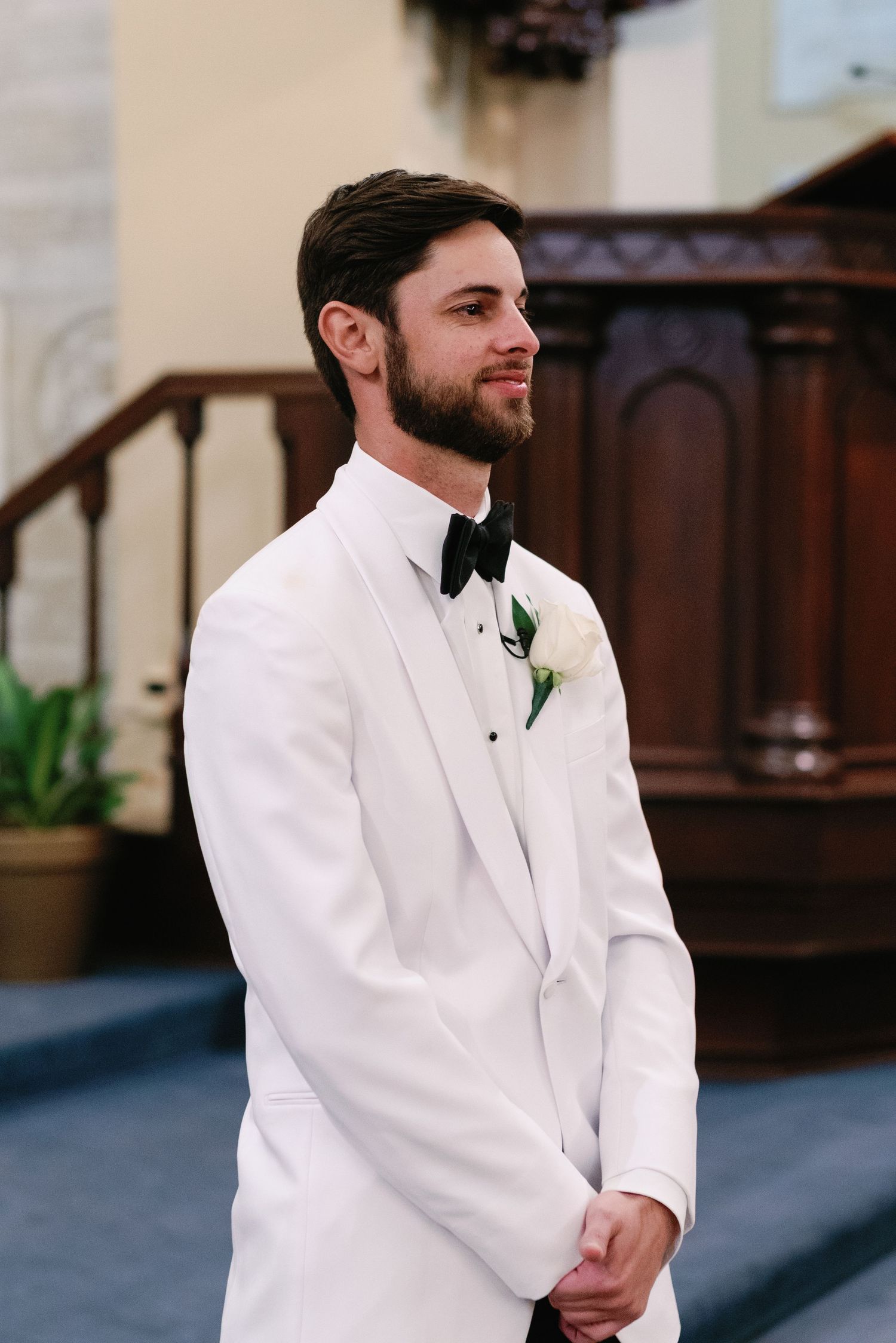 A groom in a white tuxedo jacket and black bow tie smiles while standing at the altar with a boutonniere.